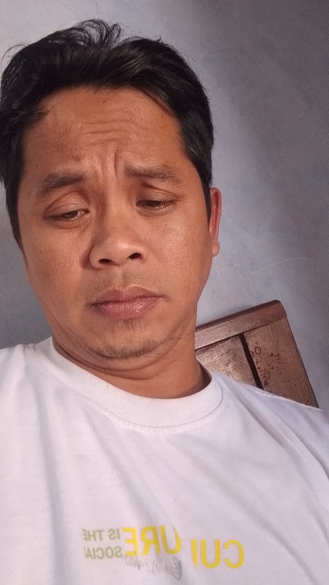 A close-up, head-and-shoulders shot depicts a middle-aged man with dark hair and brown eyes, looking downwards. He is wearing a white t-shirt with yellow lettering that reads "THE SOCIAL CUBE". The background is a plain, gray wall, with a wooden chair visible on the right side of the frame. The image appears to have been taken indoors, possibly in Karanglangu, Indonesia.