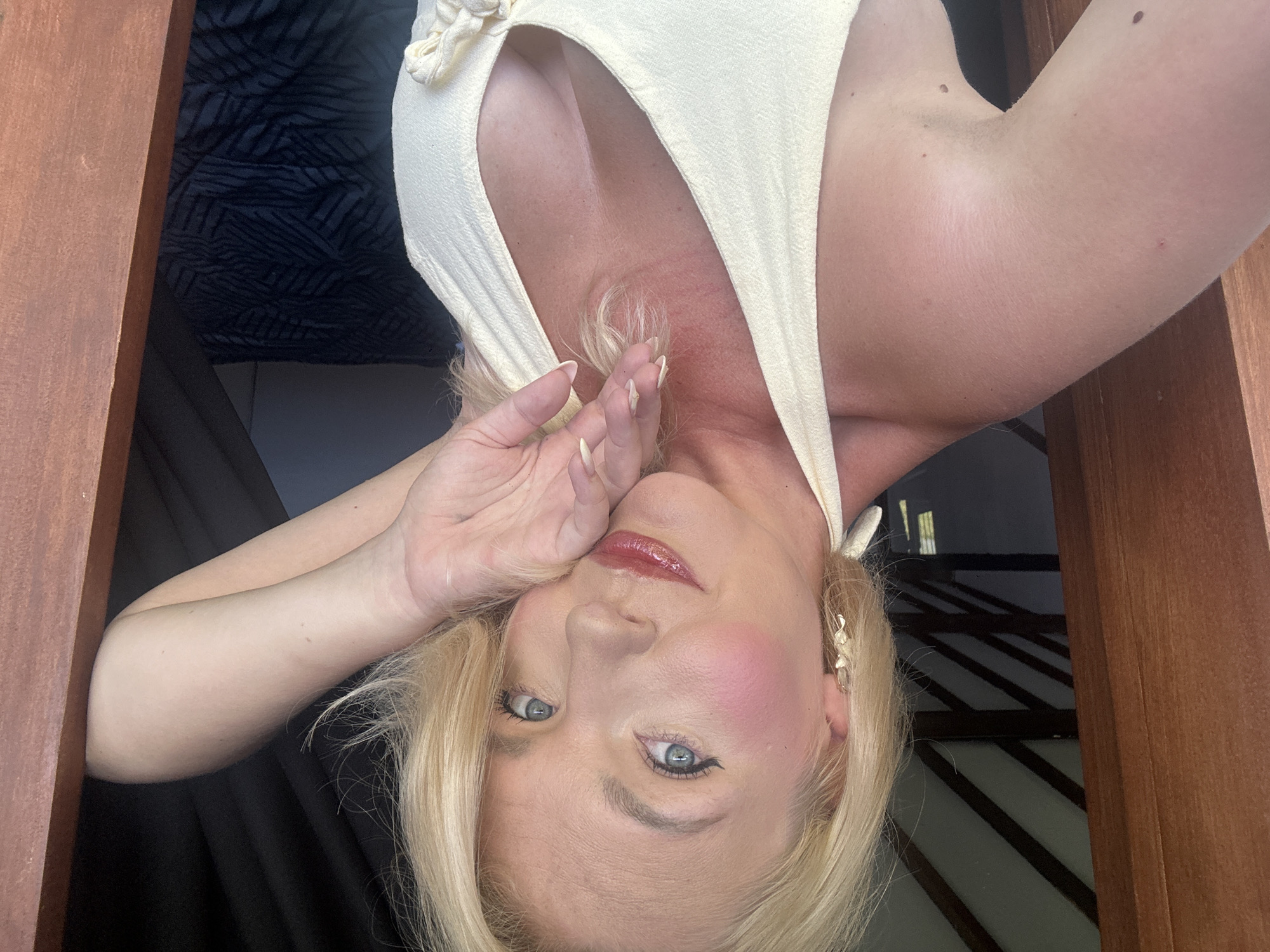 A fair-skinned blonde woman with blue eyes, wearing a light yellow halter top, leans into a wooden doorway. She has on bold pink blush, winged eyeliner, and glossy red lipstick. Her left hand rests on her cheek and her right hand plays with her hair. The background is dark, with a thatched roof visible above and a dark, patterned blanket in the background. The environment appears to be a room in a tropical location, possibly a resort in North Kuta, Indonesia.