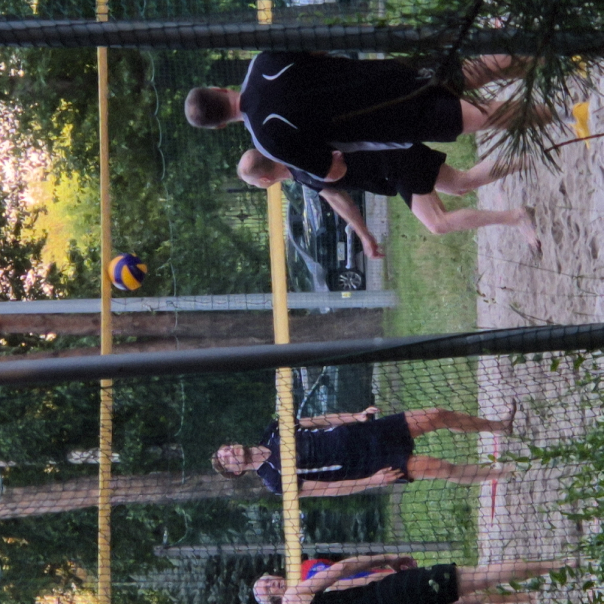 The image shows a group of people playing volleyball on a sand court. The scene appears to be outdoors, with trees and greenery visible in the background. A volleyball net is set up, and a volleyball is in play. The time of day seems to be late afternoon or early evening, as indicated by the lighting. The people are dressed in athletic wear. The setting suggests a recreational area, possibly a park or a beach. There are no visible text elements in the image. The overall atmosphere appears to be one of active recreation and friendly competition.