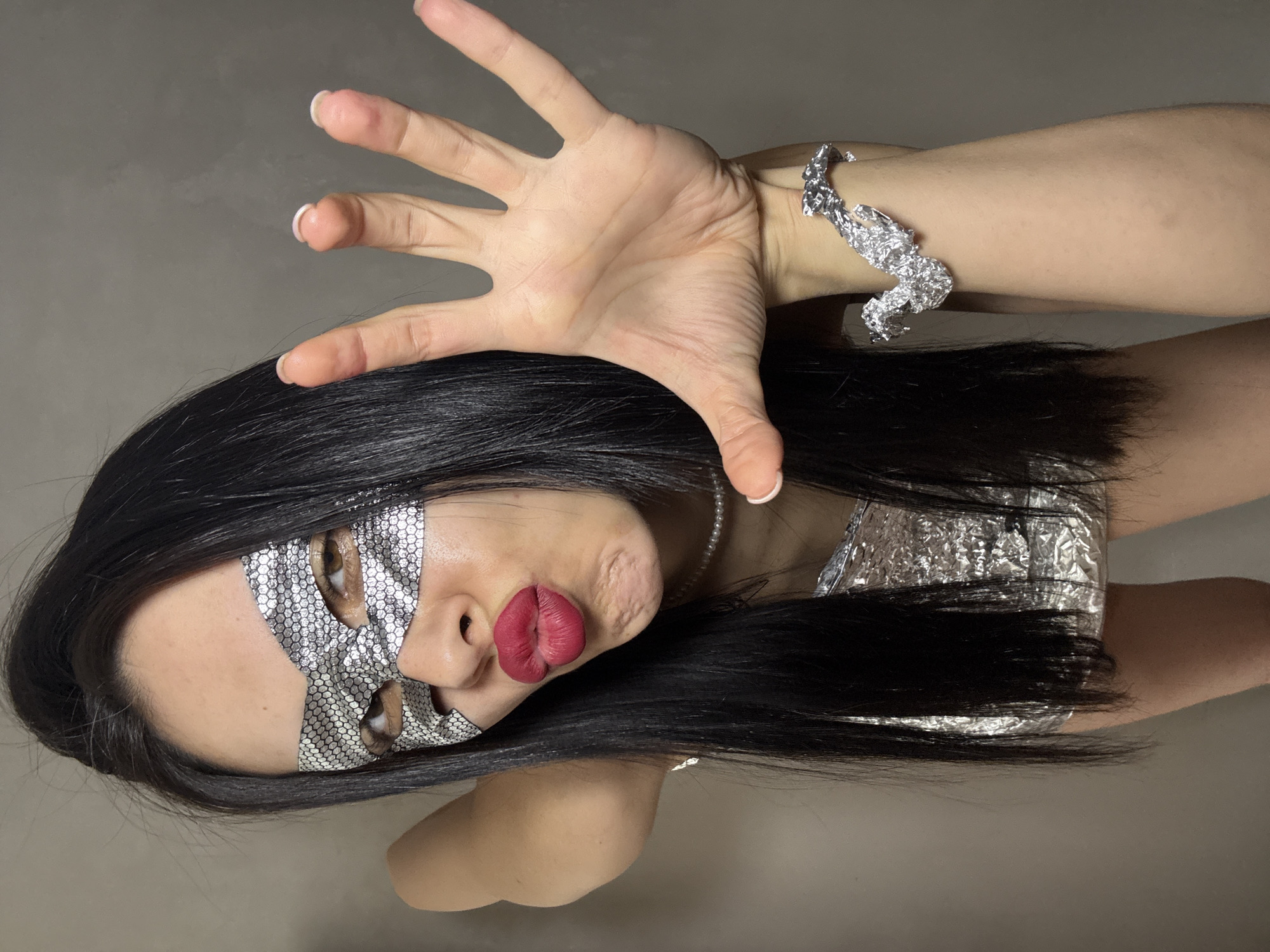 A person with long, dark hair is wearing a silver mask over their eyes and nose. They have red lipstick on and are wearing a silver bracelet on their right wrist. They are holding their right hand up with their fingers spread. The person is wearing a silver top. The background is a plain, light gray color. There is no indication of this image being in Votkinsk, Russia.