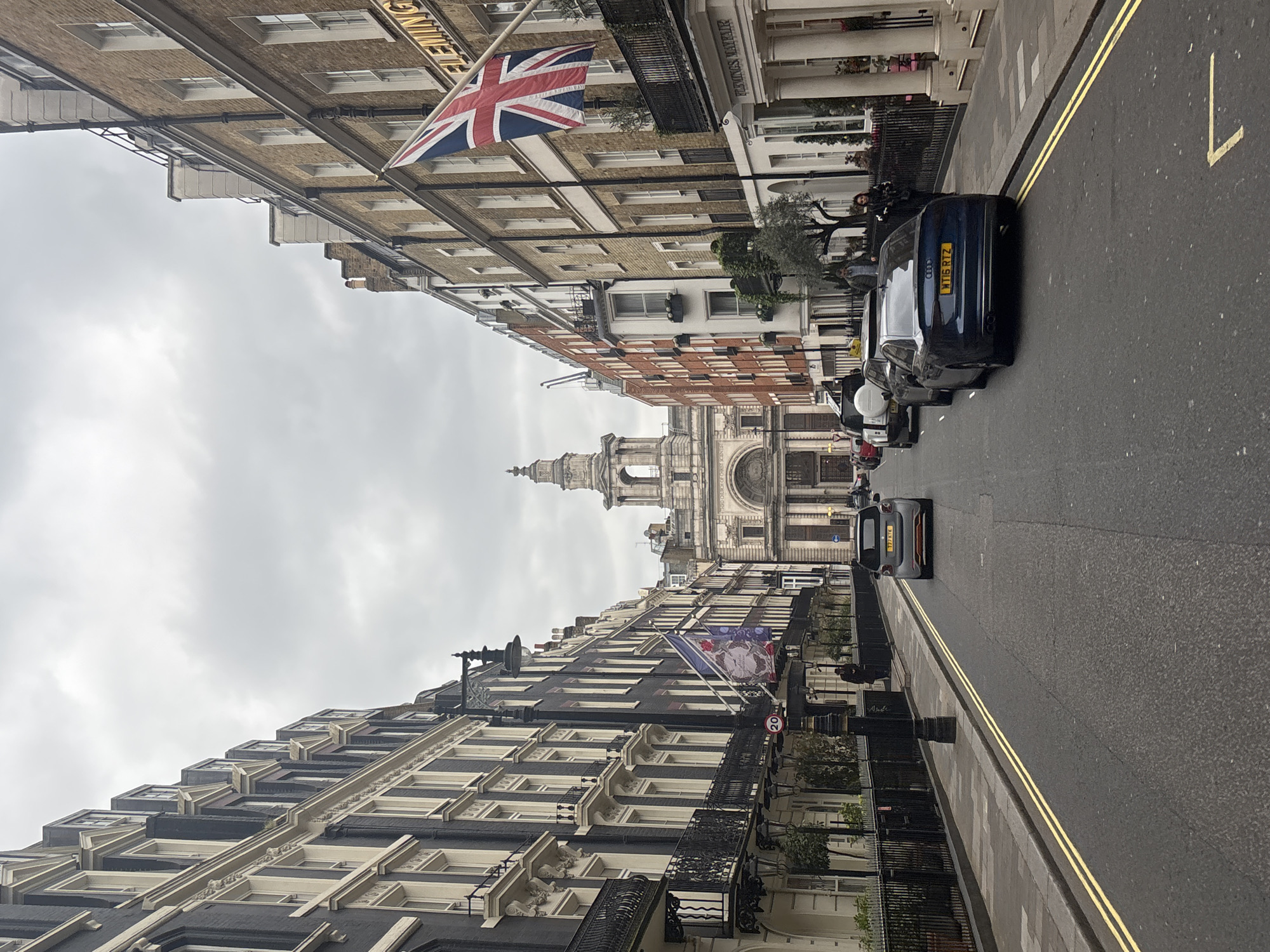 The image captures a street scene in London, United Kingdom, characterized by tall, predominantly brick and stucco buildings lining both sides of a narrow road. The sky is overcast. The Union Jack flies from a building on the right side of the street. In the distance, a building with a dome or spire can be seen. The street has double yellow lines and several cars are parked along the sides, including an Audi. A black taxi is traveling down the street towards the camera. Some pedestrians are walking on the sidewalks.