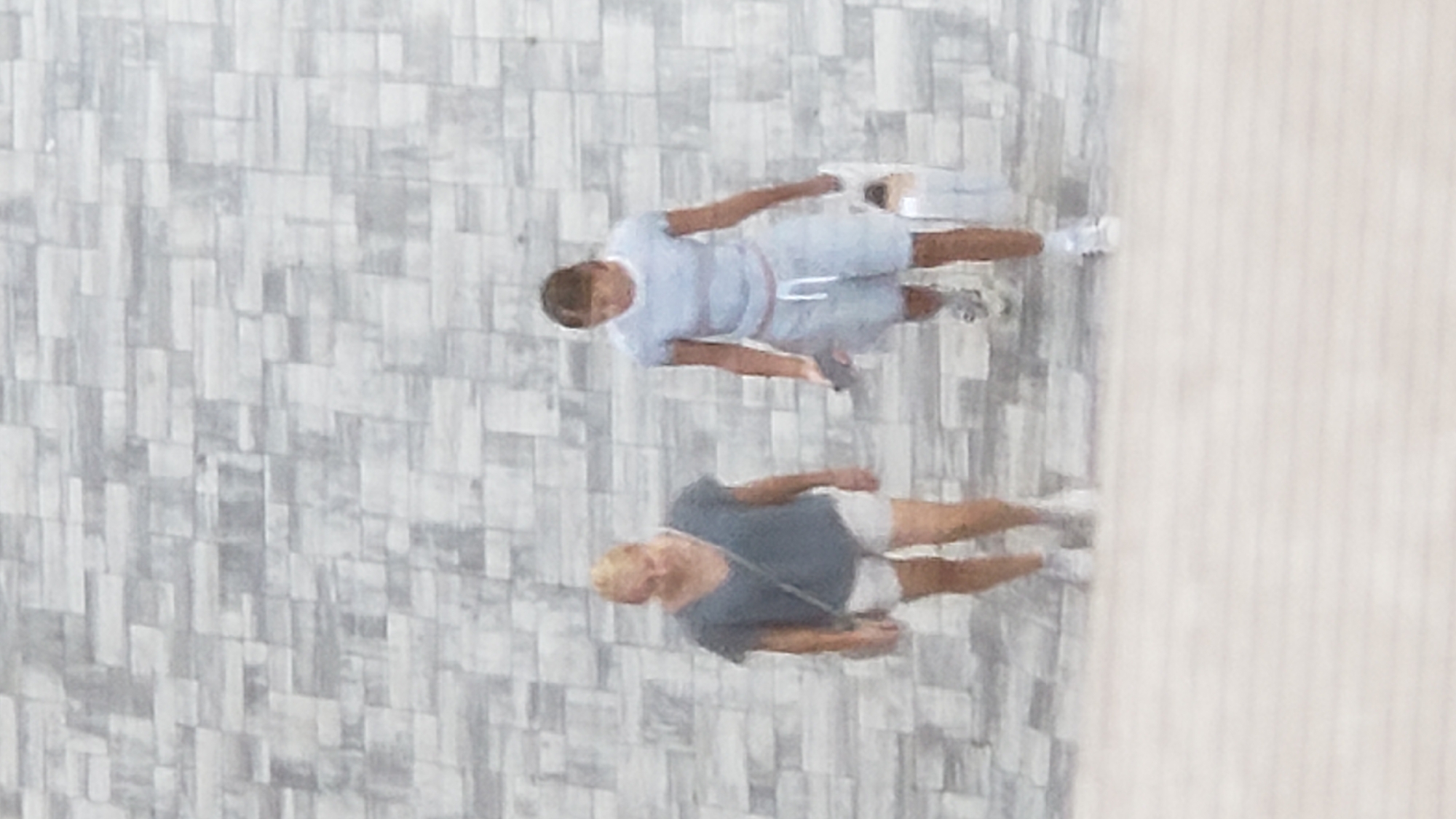 Two individuals, appearing to be women, walk on a patterned stone surface, likely a sidewalk or pathway in a city. The woman on the left wears a grey top and white shorts. The woman on the right wears a light blue t-shirt and light blue shorts. The background consists of a textured grey stone wall. The scene is outdoors, and the lighting suggests daytime. The image quality is low, with a blurred effect. Without specific landmarks or identifying features, the location cannot be definitively determined as Prešov, Slovakia, or any other specific city.
