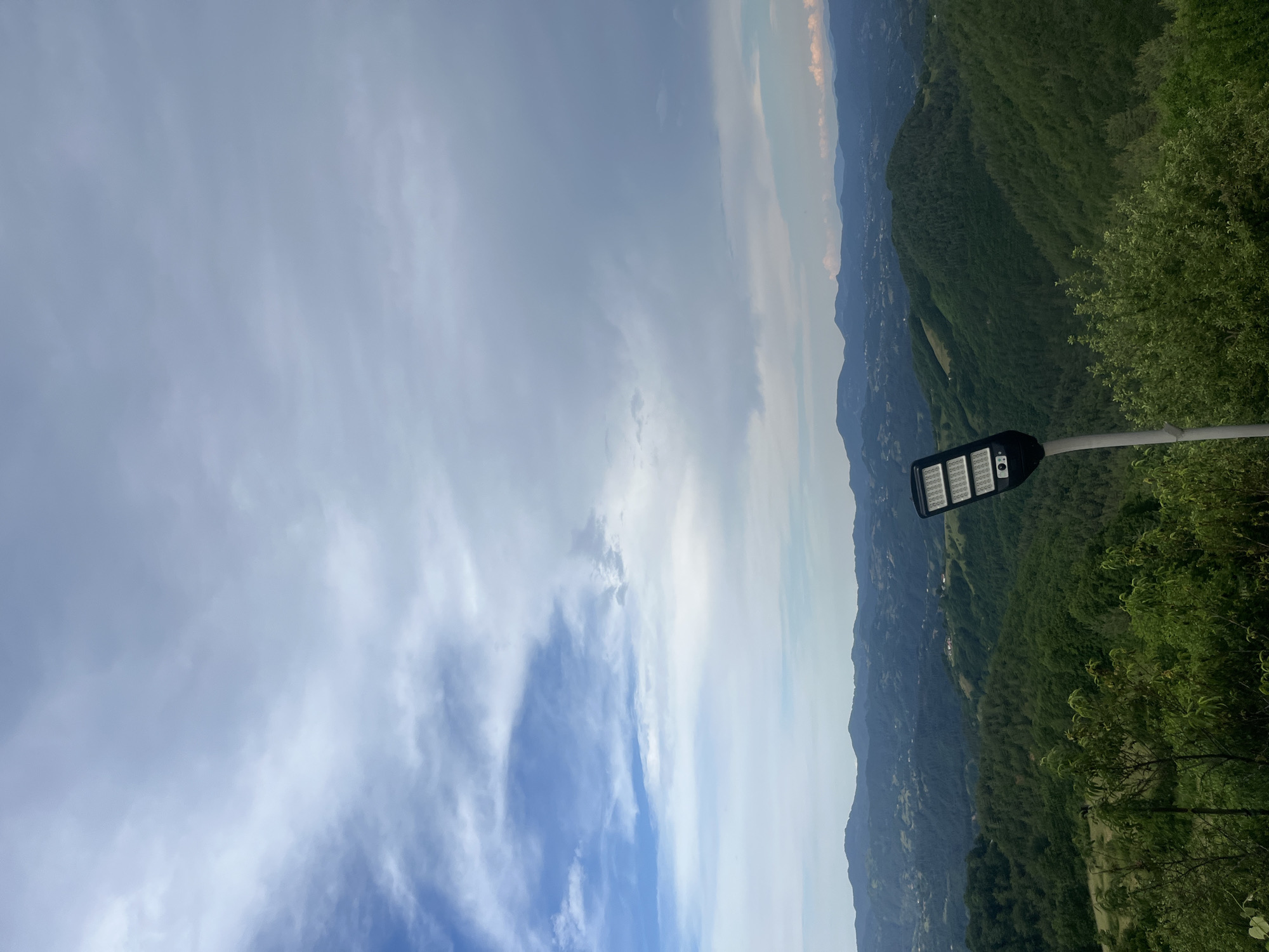 A street lamp with a black rectangular fixture and a white pole stands in the foreground against a backdrop of rolling, verdant hills and a cloudy sky. The lamp is positioned slightly to the right of the frame's center, with its illuminated panel facing the viewer. The hills are covered in dense green vegetation, with some visible structures and roads scattered throughout. In the distance, more layers of hills fade into a hazy horizon. The sky is a mix of blue patches and large, diffused clouds. The overall scene conveys a sense of a rural or semi-rural mountainous landscape under an overcast sky.