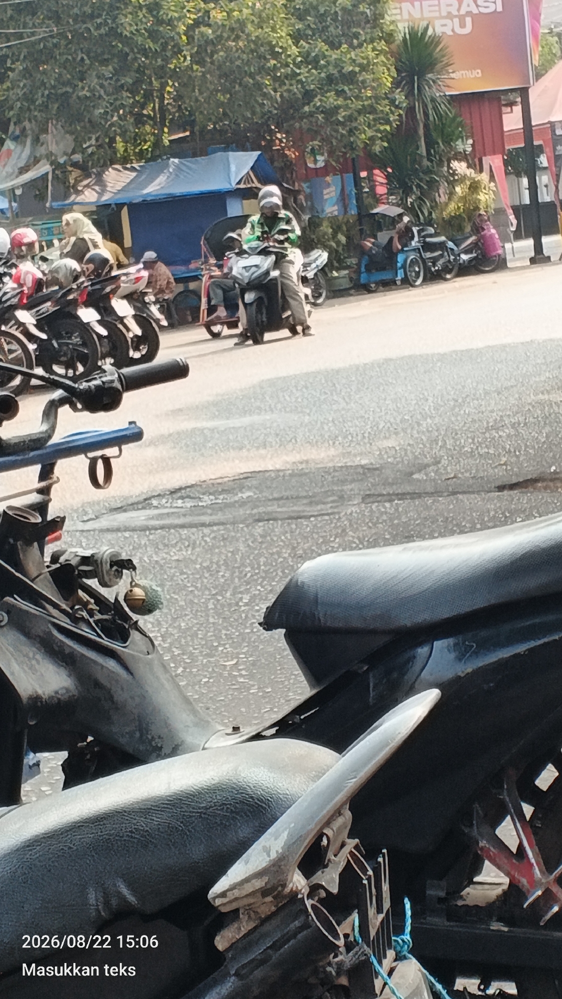 A sunny, outdoor daytime scene in Malang, Indonesia, features a street with several motorcycles and a few people. In the foreground, parts of a black motorcycle are visible, including its seat and handlebars. A blue frame with a ring is attached to the handlebars. In the middle ground, a man wearing a green jacket and a striped helmet rides a motorcycle. To his left, another motorcycle is parked, and beyond that, a blue tarp-covered structure is visible. Further back, a large billboard with the text "GENERASI BARU" and a stylized image of a globe is prominent. The background shows trees and more buildings. The road surface is asphalt. The timestamp "2026/08/22 15:06" and the text "Masukkan teks" are overlaid on the lower left of the image.
