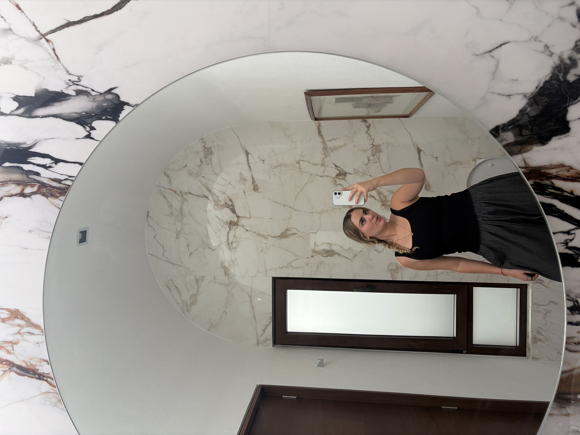 A woman is captured in a mirror selfie, holding a white smartphone with her right hand to take the picture. She has blonde hair styled in a braid, and is wearing a black tank top and a dark, possibly pleated, skirt or wide-leg pants. Her expression is neutral as she focuses on her phone.
The image is taken in an indoor setting, likely a modern bathroom or hallway in San Bernardino Tlaxcalancingo, Mexico. A large, circular or oval mirror occupies the central part of the frame. The wall behind the mirror is adorned with a distinct white marble pattern featuring prominent black and brown veins. Inside the mirror's reflection, the room appears to have lighter, white marble walls with subtle brown veining.
Visible within the reflection are a dark brown wooden window frame with an opaque or frosted glass pane, a smaller, similar window section beside it, and part of a dark brown wooden door at the bottom of the reflection. Above the larger window in the reflection, a framed picture is partially visible on the wall. The wall surrounding the mirror and visible outside its reflection also features the same white marble with black and brown veins, indicating continuity in the interior design. The lighting suggests daytime, with diffused light coming through the window. There is no visible text in the image.