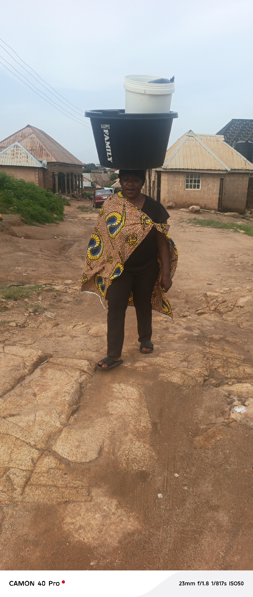 A woman in Nigeria is captured in a full-length, eye-level shot, carrying two buckets on her head. The larger, black bucket is on the bottom, with "HAPPY FAMILY" written vertically on its side. A smaller white bucket is stacked on top of the black one. The woman is wearing dark pants, a black t-shirt, and a colorful patterned wrap draped over her shoulders. She has black hair and appears to be walking on a dirt path with large rocks.
In the background, there are several small, simple structures that appear to be houses, some with corrugated metal roofs and others with tiled roofs. There's also a red car visible in the distance. The setting suggests a rural or semi-rural environment in Nigeria. The sky is overcast, indicating a cloudy day. The woman's expression is neutral, and the overall mood of the image is one of everyday life and labor. The timestamp at the bottom reads "23mm f/1.8 1/817s ISO50". Another text reads "CAMON 40 Pro".