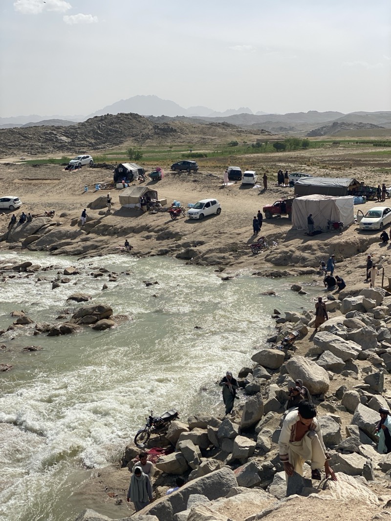 The image depicts a riverine landscape with a congregation of people, vehicles, and tents. The river, characterized by its rocky bed and turbulent flow, forms a prominent part of the foreground. People are situated both along the riverbank, climbing on rocks, and further inland, near makeshift encampments.
A mix of vehicles, including white cars and a red truck, are parked on the terrain away from the river's edge. Tents of various sizes are erected nearby, suggesting a temporary settlement or gathering. The background shows a landscape featuring elevated terrain and a somewhat hazy sky.
The presence of people engaging in various activities near the river and the setup of tents indicates this is a populated area. The location is identified as Kim, Afghanistan.