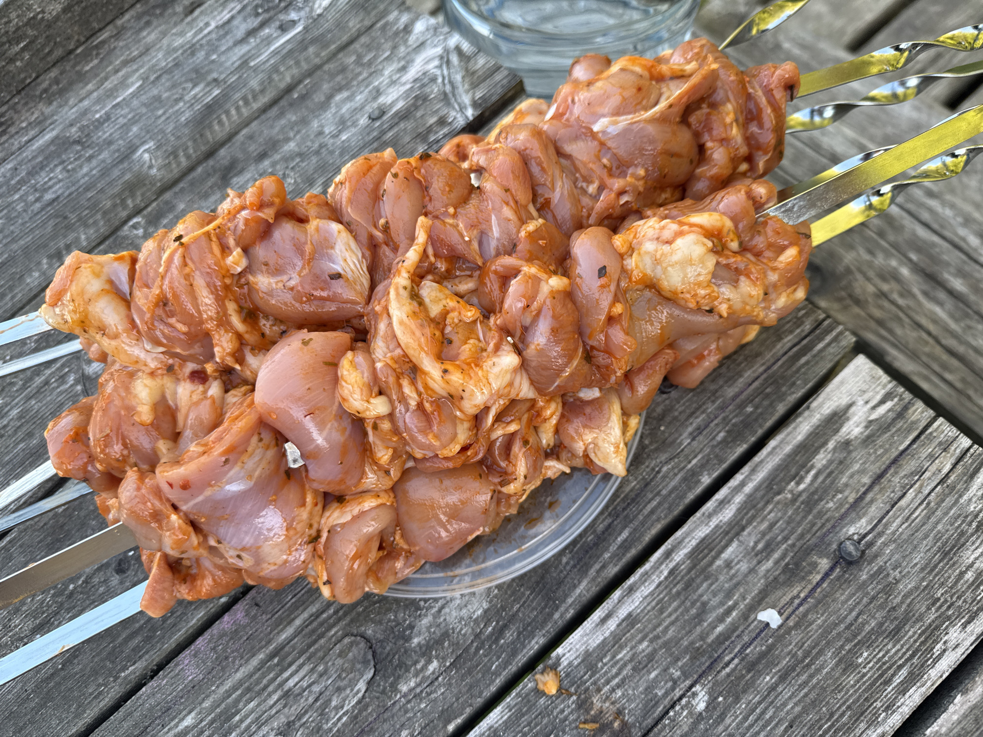 A top-down, medium shot captures a close-up of raw chicken skewers on a weathered wooden table. The chicken pieces are cut into bite-sized chunks, marinated in a reddish-orange sauce, and threaded onto metal skewers. The skewers are arranged diagonally across the frame, with the handles extending out of view to the right. The wooden table has a visible grain and appears to be outdoors, suggested by the natural lighting. To the right of the skewers, a portion of a clear glass object, possibly a pitcher or bottle, is visible. The overall scene depicts food preparation, likely for grilling. The provided location of Zamosh'e (Zaklinskoe S/P), Russia, offers geographical context. There are no people or animals present in the image.