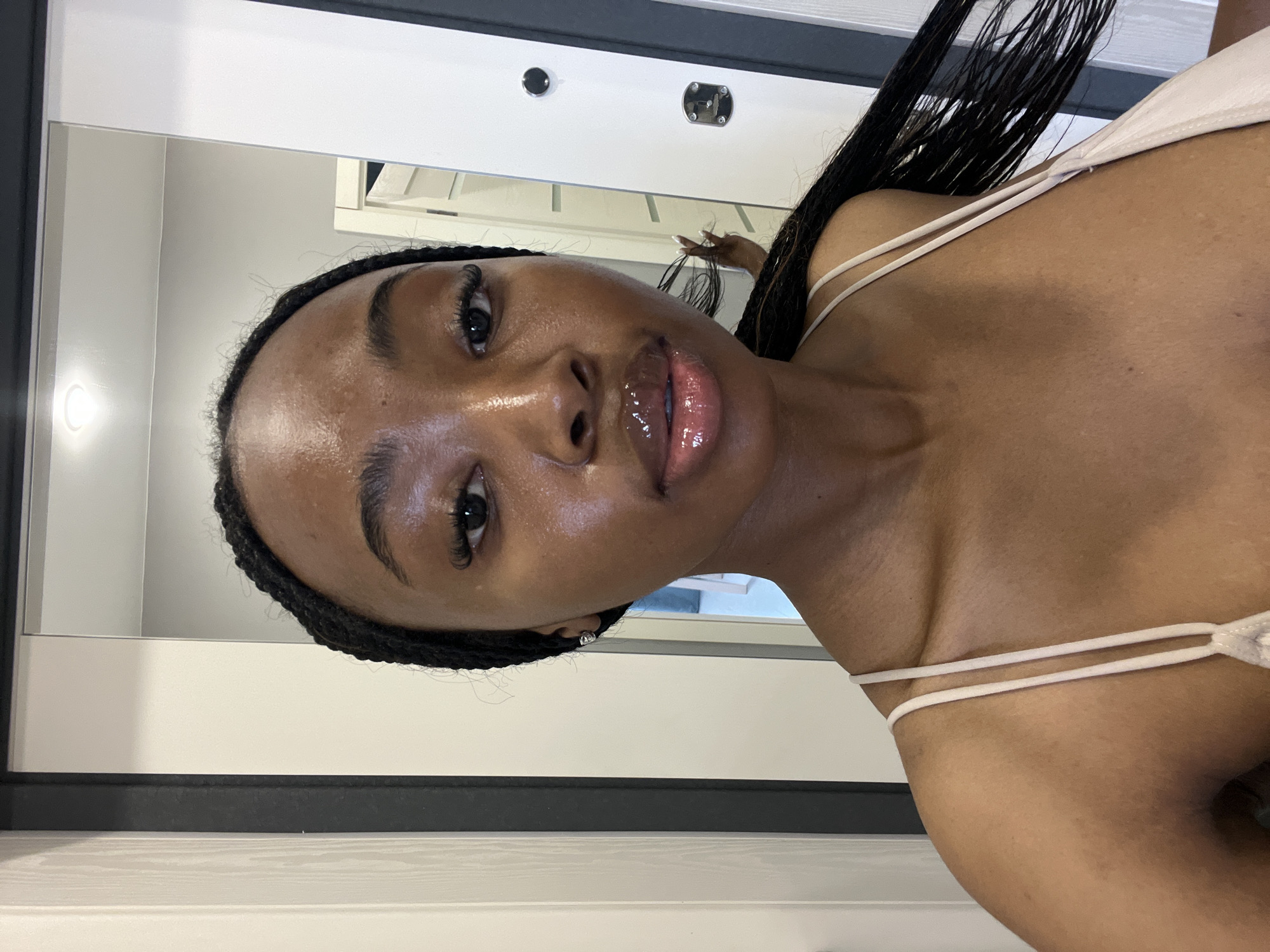 A young Black woman with braided hair and shiny lips is looking into a mirror. She is wearing a light-colored camisole. The background appears to be an indoor setting with a closet door and part of another door visible. There is a reflection in the mirror of the woman's arm and hand. The location is not specified as Kazan, Russia, or any other city.
