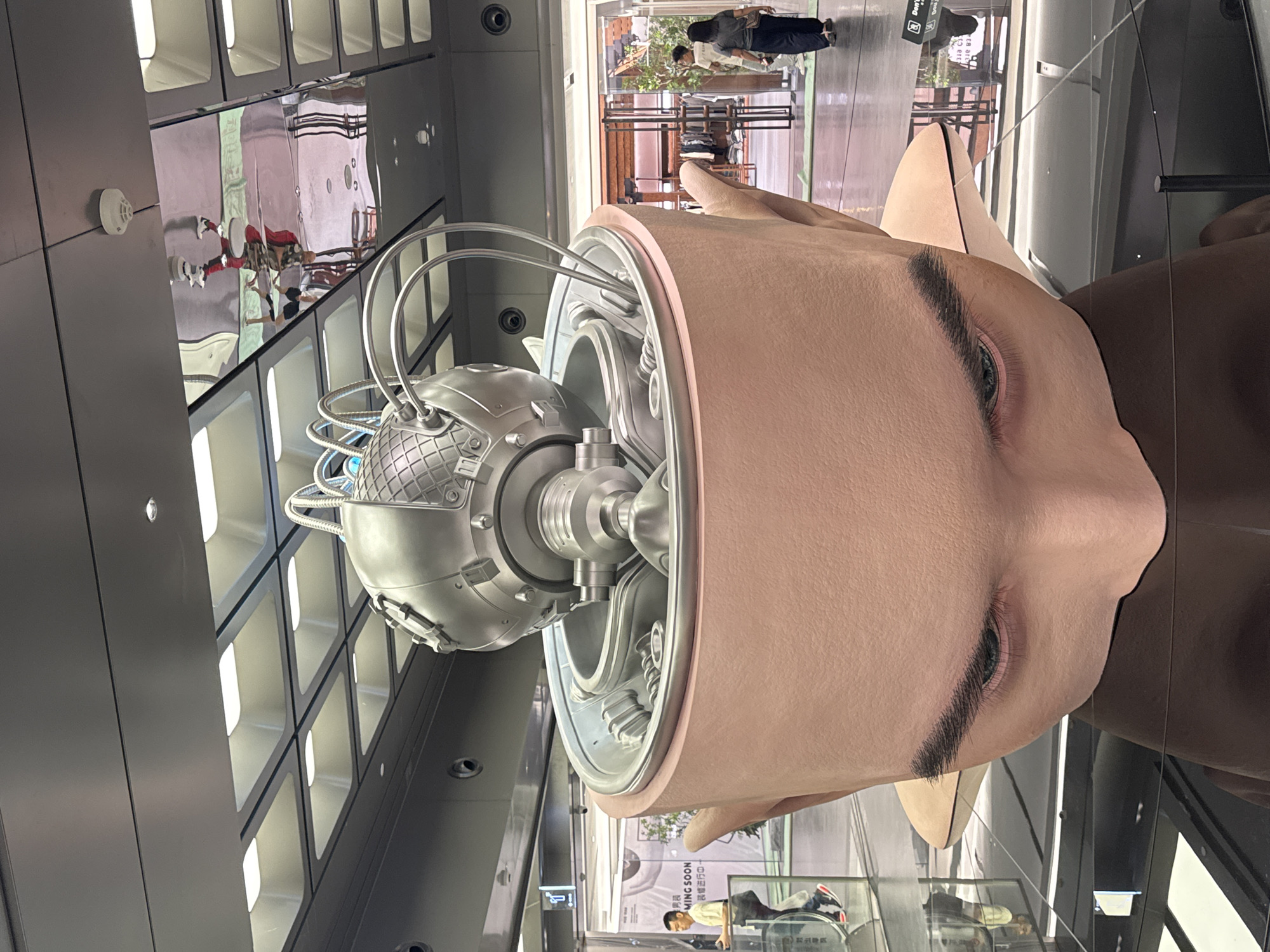 The image shows a large-scale art installation of an open human head with a metallic, technological-looking sphere exposed within. The head is humanoid in appearance, with visible facial features such as eyes and eyebrows. The interior sphere is connected to the head by cables and mechanical components.
The artwork is displayed within a modern, multi-story building with a grid-like facade. Reflections in the glass surfaces of the building show other people and interior spaces. The location is the city of Liwan District, China.