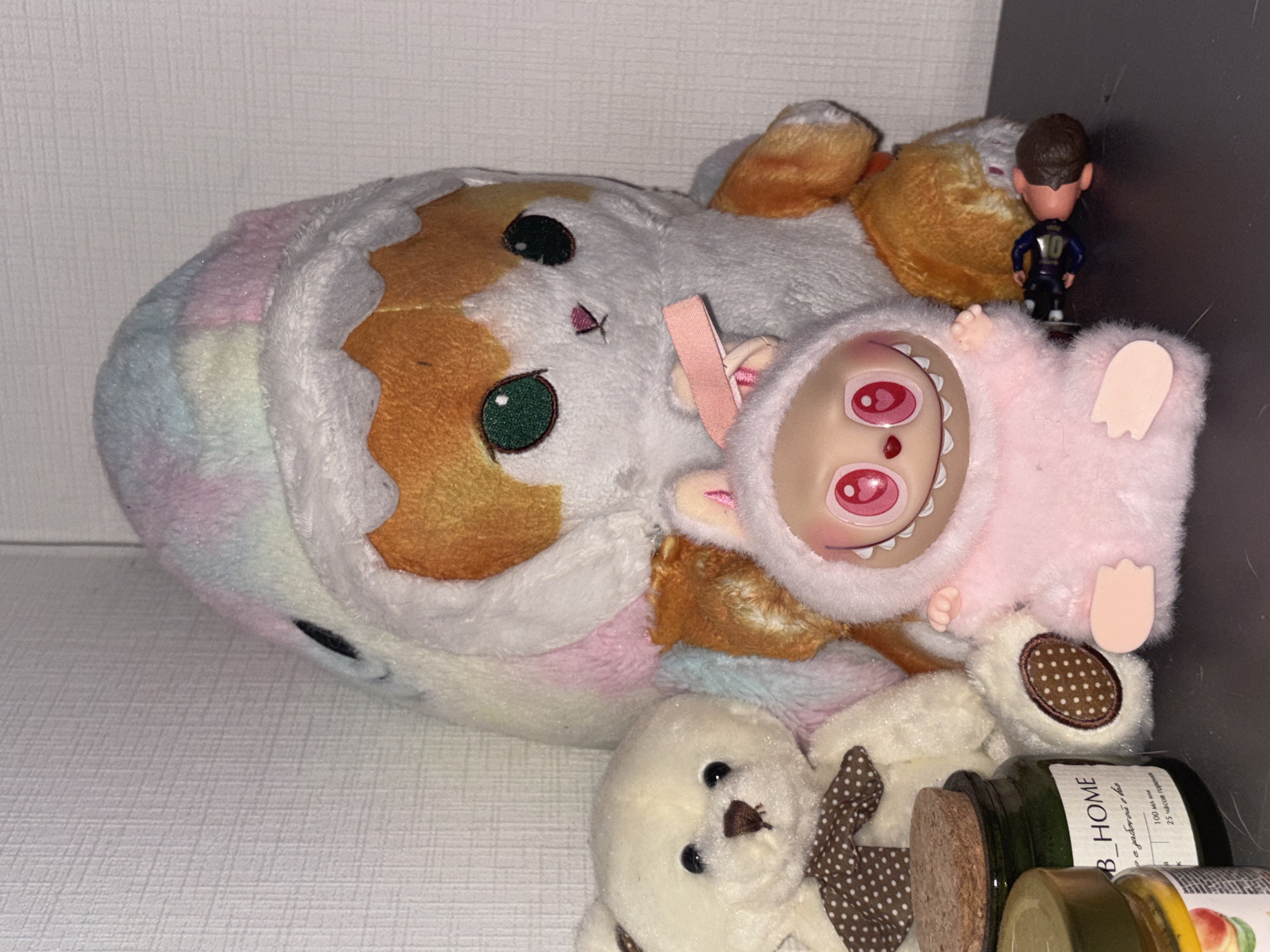 The image depicts a shelf cluttered with various objects. Prominently featured are several plush toys, including a rainbow-colored cat, a pink furry doll with a wide-eyed expression, and a small white teddy bear with a polka-dot bow tie. Adjacent to the plush toys are several cylindrical containers, appearing to be jars or bottles, one labeled "B_HOME". A small figurine of a soccer player is also visible in the background. The environment appears to be an interior shelf within a home. The specific location is unknown and cannot be determined from the image.
