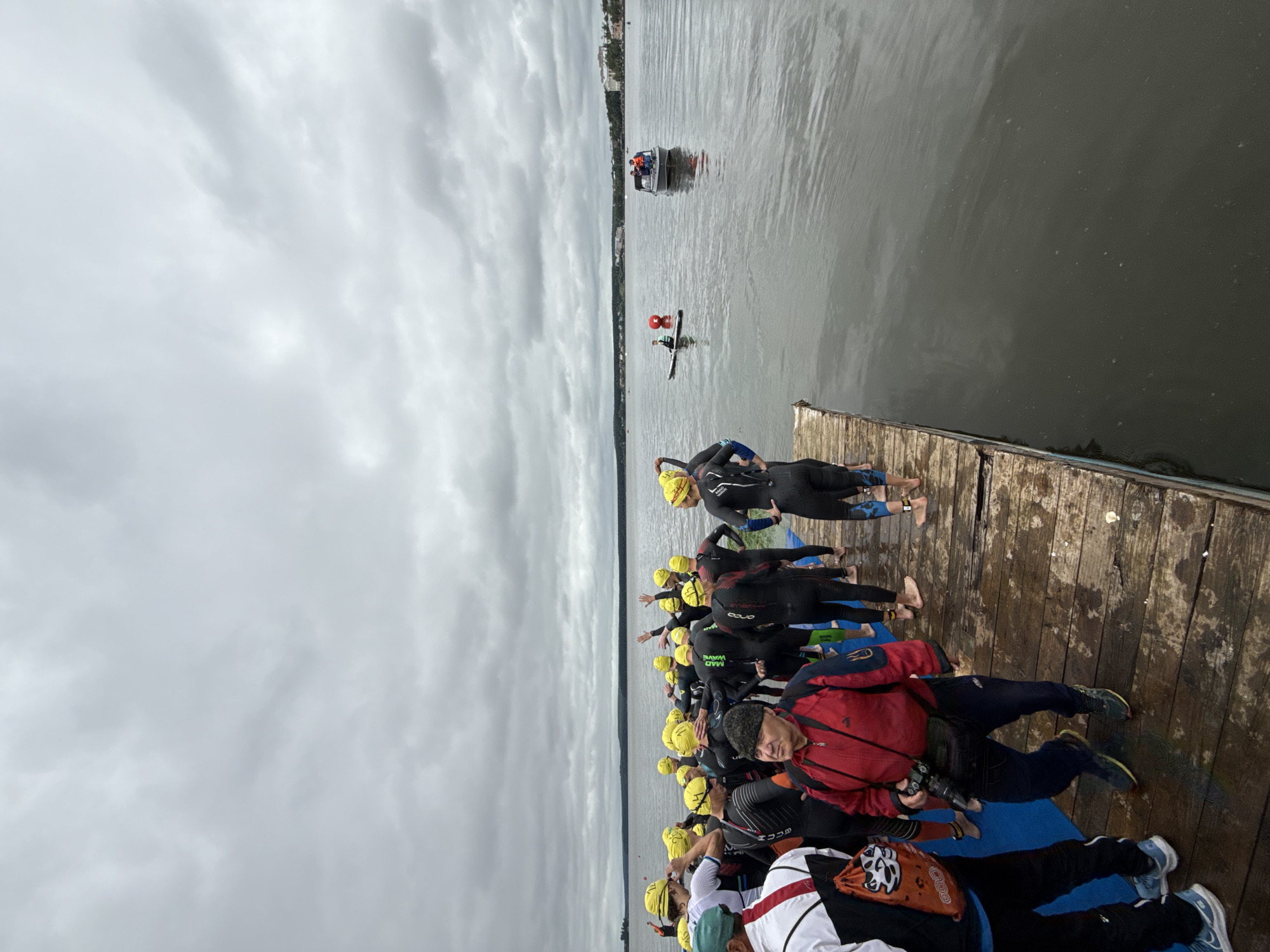 A group of people in yellow swim caps and wetsuits are gathered on a wooden dock, preparing for a swim. The sky is overcast, and the water is calm. In the distance, a few buildings are visible along the shoreline. A small boat with several people on board is visible on the water, as is a person on a paddleboard. The scene appears to be at the Votkinsk Reservoir in Votkinsk, Russia.