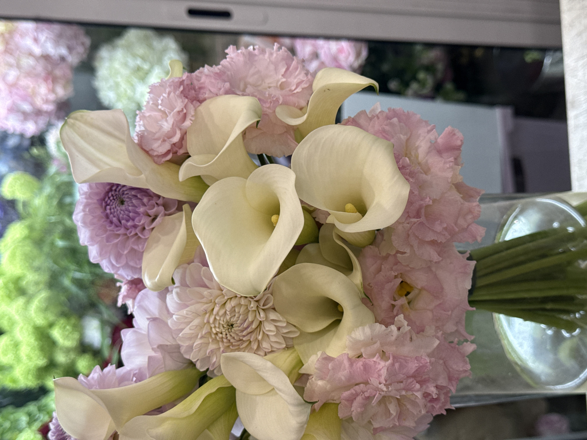 A close-up, eye-level shot captures a bouquet of pale yellow calla lilies and soft pink carnations arranged in a clear glass vase. The flowers are clustered together, with the calla lilies' trumpet-shaped blooms prominent in the foreground. In the background, out-of-focus green foliage and pastel-colored flowers, possibly hydrangeas, create a soft, blurred backdrop. The image is well-lit, with natural light enhancing the delicate textures and colors of the floral arrangement. The scene appears to be indoors, possibly in a flower shop or home setting. The presence of a light-colored surface in the foreground suggests a table or counter. The image does not depict any people or identifiable landmarks, and therefore, its location within Assinovskaya, Russia, cannot be determined from the visual information provided.