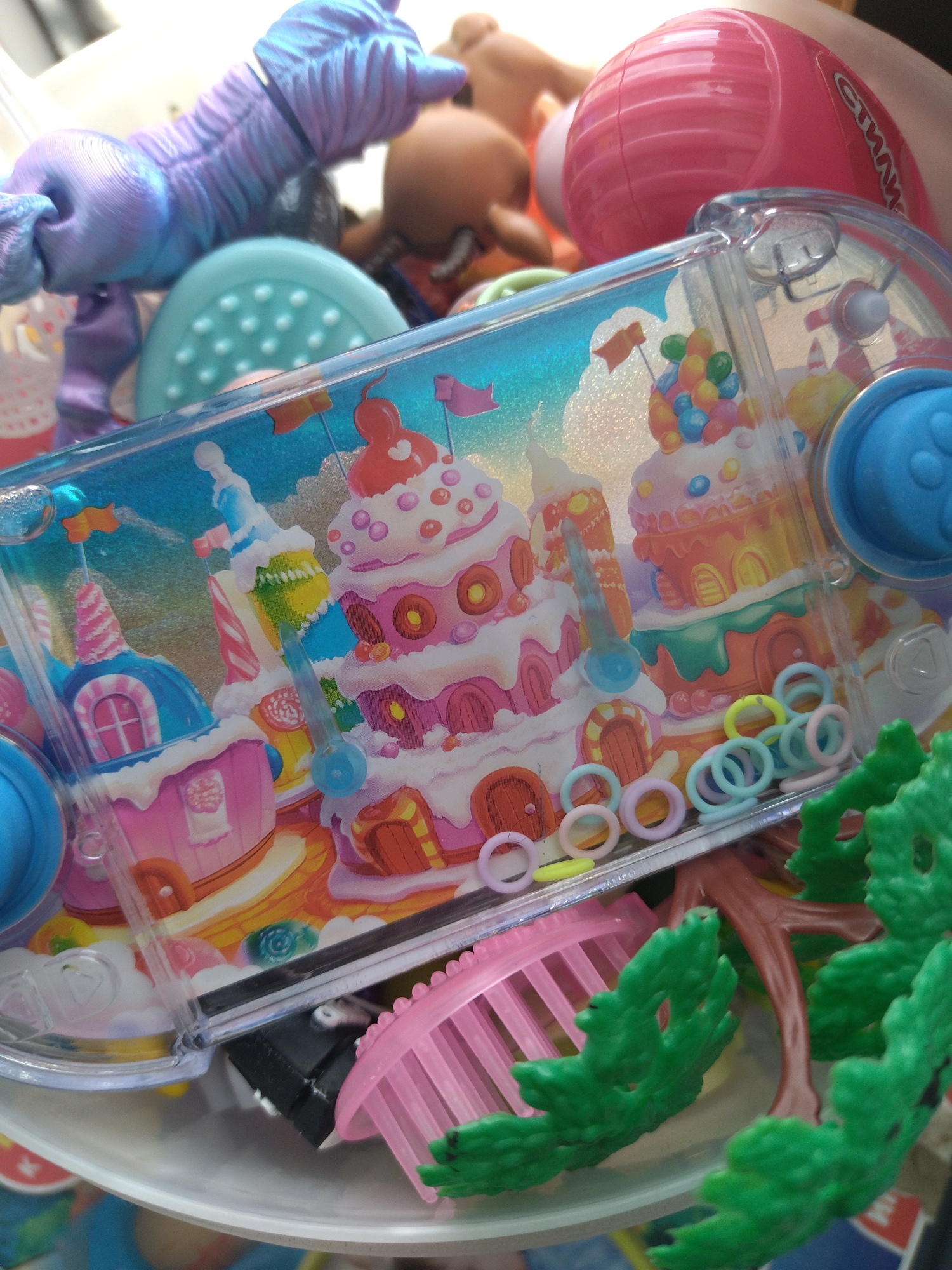 This is a close-up, high-angle, eye-level shot of a collection of children's toys. The dominant subject is a handheld water game with a colorful, cartoonish design featuring candy and ice cream-themed castles. The game is partially submerged in a white plastic container, and other toys are visible around it.
In the background, a purple, textured toy with jointed limbs, possibly a doll or action figure, is visible on the left. To its right, a light blue plastic dome with raised dots is partially obscured. Further back, a tan-colored toy head with a bald scalp and a simplified facial structure is visible. On the far right, a shiny pink spherical object with horizontal ridges and some text on it, possibly a ball or part of another toy, is prominent.
In the foreground, several small, colorful plastic rings in pastel shades of pink, blue, and yellow are scattered across the water game's surface and around it. A pink, comb-like plastic object with rounded teeth is positioned below the water game, and to its right, a brown, tree-like plastic structure with green leafy elements is visible. The overall environment appears to be indoors, with soft, diffused lighting. There are no discernible landmarks or elements that would suggest a specific location within Izhevsk, Russia.