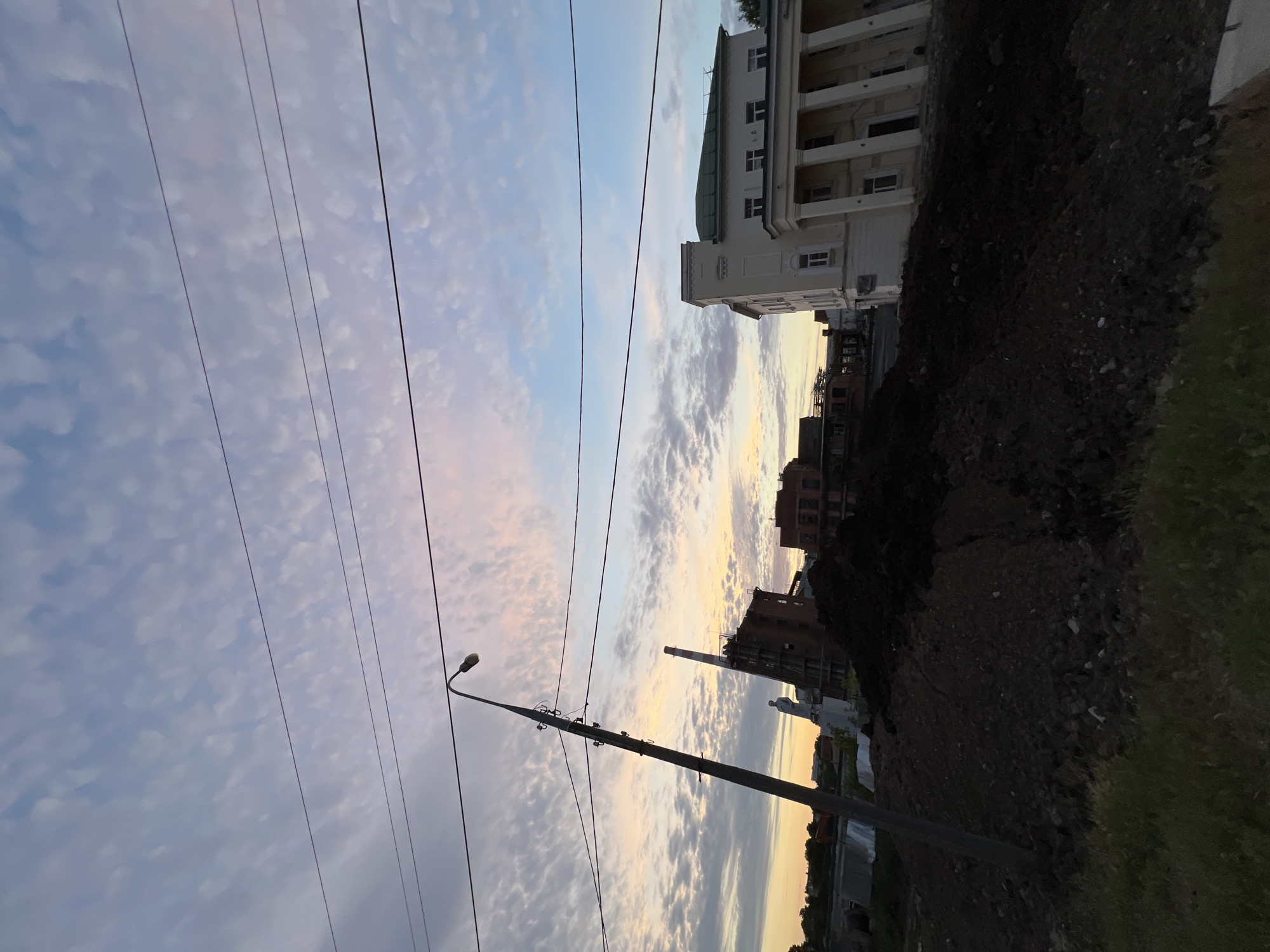 This media file captures a view of buildings and a sky filled with clouds, likely taken in Krasnodar, Russia. The perspective is tilted, suggesting the photograph was taken from a side angle, perhaps from a street.
The subjects include several buildings of varying architectural styles, one of which appears to be a larger, more ornate structure with columns and multiple windows. There are also smaller, brick buildings visible. Utility poles with wires crisscross the frame, indicating an urban or suburban setting. A street lamp is also present. The ground in the foreground is dark and appears to be covered in dirt and rocks, with a patch of green grass to the right.
The time of day is likely late afternoon or early evening, as indicated by the soft, diffused light and the pastel colors in the sky, which feature a mix of blues, pinks, and yellows. The clouds are scattered and appear wispy. The weather seems to be clear, with no obvious signs of rain or strong winds.
There is no visible text in the media file. The overall mood conveyed is one of a calm, transitional moment, possibly a quiet evening in an urban environment.