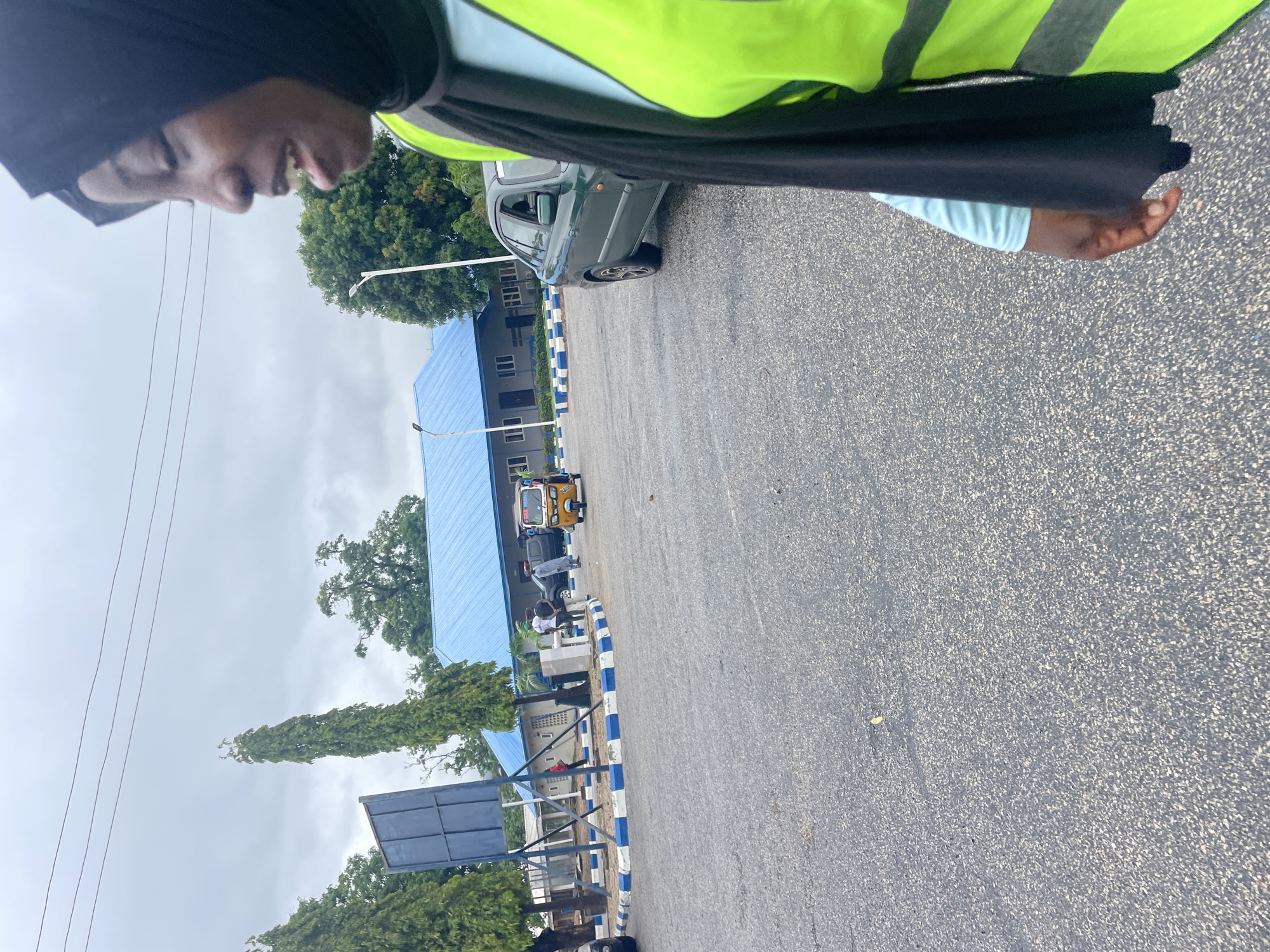 In Lokoja, Nigeria, a medium shot captures a side view of a woman wearing a black hijab and a bright yellow-green safety vest over a light blue long-sleeved shirt. She is looking to her right with her mouth open, as if speaking or laughing. In the background, a blue-roofed building with white window frames and a darker grey facade stands under an overcast sky. A dark green sedan is parked in front of the building to the right of a yellow auto-rickshaw. Several individuals are standing near the building and the vehicles, and a tall, narrow evergreen tree is positioned to the left of the building. The foreground consists of an asphalt road surface with a distinct white and blue striped curb line.