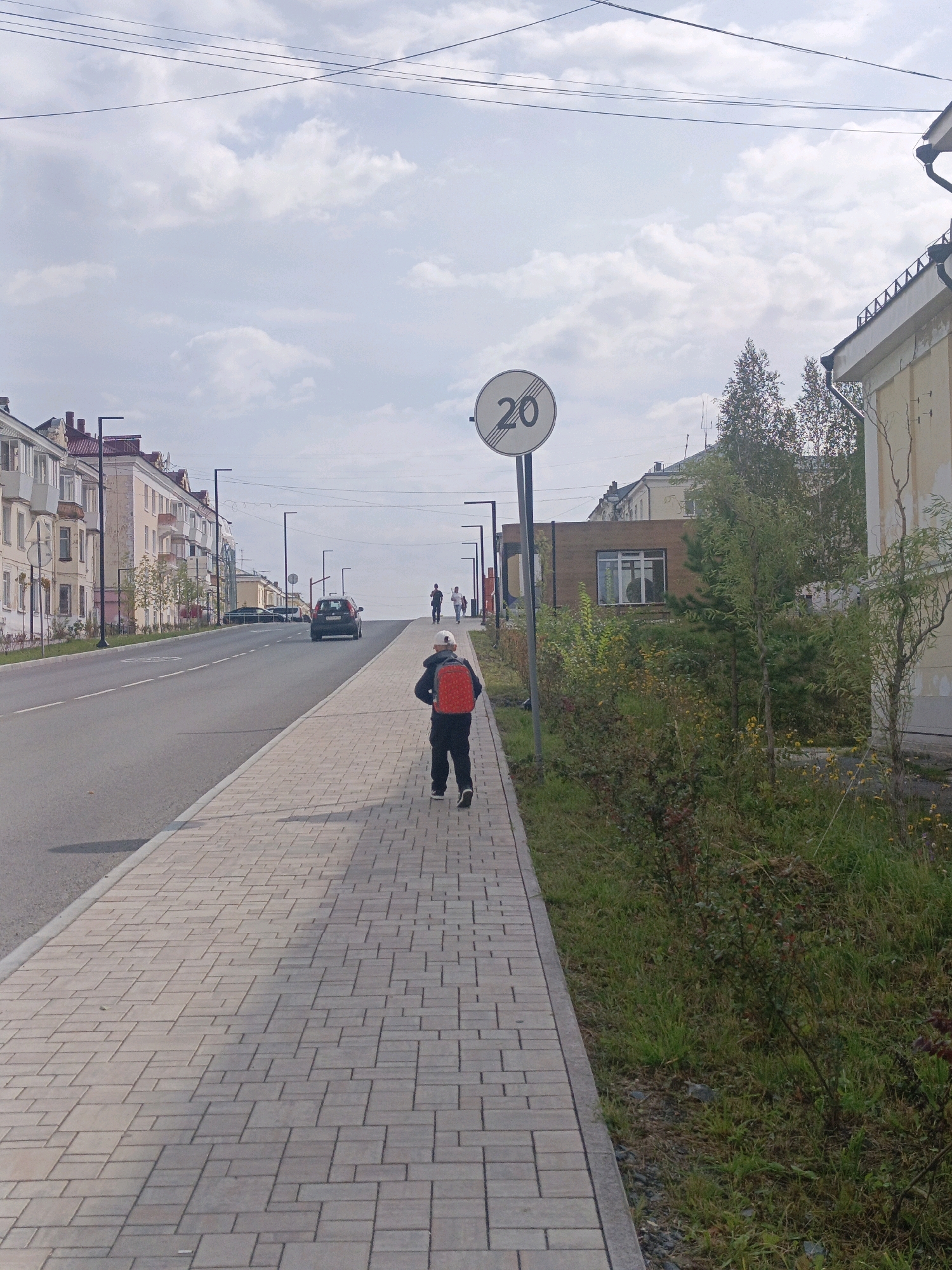 The image depicts a street scene in the city of Satka, Russia, likely on a cloudy day. In the foreground, a young person wearing a red backpack walks away from the camera on a paved sidewalk. The sidewalk is constructed of light-colored rectangular pavers laid in a staggered pattern. Adjacent to the sidewalk is a strip of grass and low-lying bushes.
The street itself is paved with asphalt and slopes slightly uphill. There are at least two lanes of traffic visible. Several cars are present on the road, including a dark-colored sedan in the middle distance. In the background, there are residential buildings with multiple stories, characterized by light-colored facades and dark roofs. Streetlights are visible along the sidewalk.
A round traffic sign indicating a speed limit of 20 is posted on a pole to the left of the person. In the far background, more buildings and trees are visible, and the sky is overcast with some patches of blue. The overall environment suggests a suburban or urban residential area.