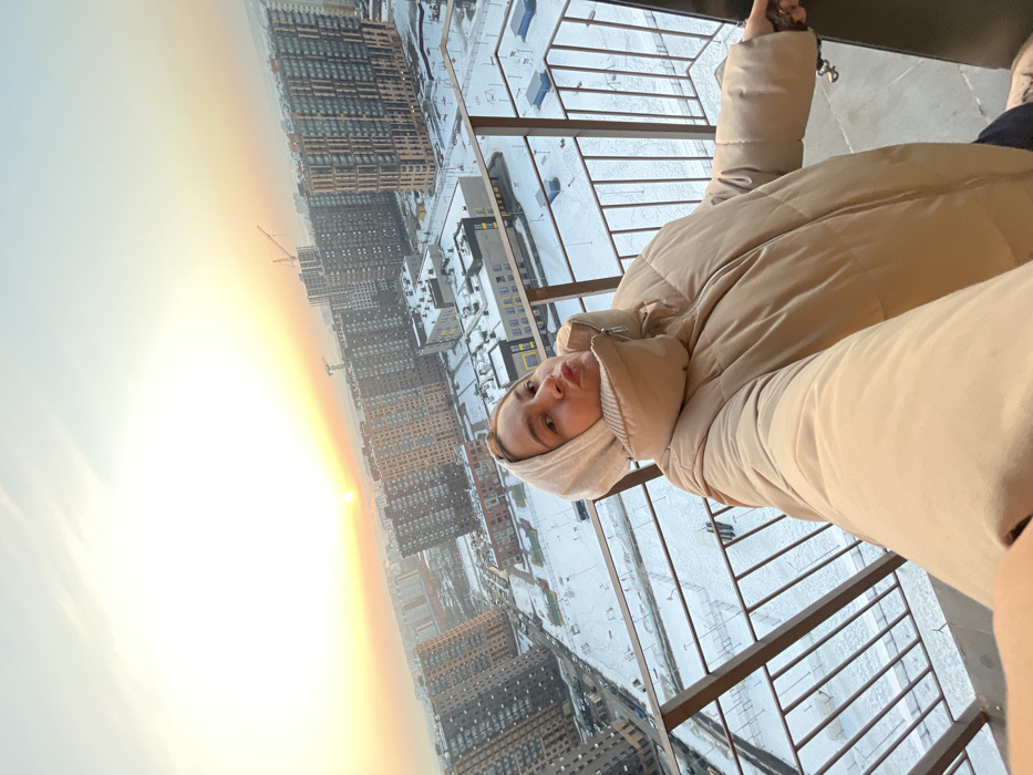A female individual, approximately 20-35 years old, is depicted in a selfie taken on an elevated outdoor balcony in Elektrougli, Russia. She is wearing a light beige hooded puffer jacket, with a beige fabric element covering part of her head and neck. Her lips are pursed, and her eyes are directed towards the camera. Her right hand is extended, presumably holding the camera device, while her left arm rests against a dark brown metal balcony railing.
The background presents an expansive cityscape under winter conditions, with all visible ground surfaces covered in snow. Numerous multi-story residential buildings of varying modern architectural styles, predominantly in brown, grey, and white tones, populate the urban landscape. Several construction cranes are visible in the distance among the buildings, indicating ongoing development. Lower institutional-type buildings are also scattered throughout the scene. The sky occupies the upper frame, transitioning from a bright, intense golden-orange hue on the left horizon, where the sun is visible as a distinct disc, to a lighter blue-grey overhead. The low position of the sun suggests either sunrise or sunset.
