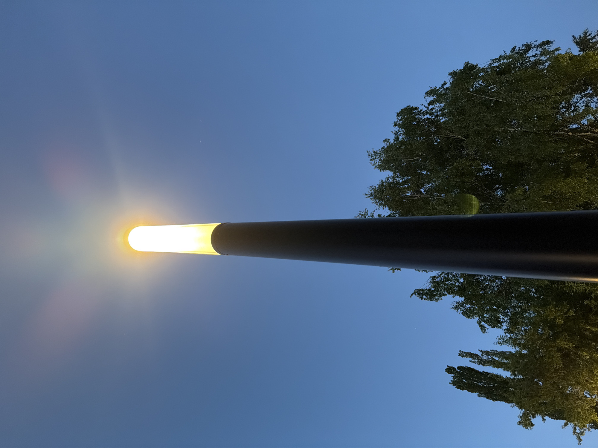 The image shows a street lamp at night, viewed from below, pointed vertically upwards. The lamp has a long, cylindrical shape and emits a bright, yellow light. The pole is a dark color and narrows towards the light source. The sky is a gradient of deep blue to lighter blue as it approaches the light source. The upper right part of the image shows a tree with green foliage. There are no people in the image. The location appears to be an urban environment.