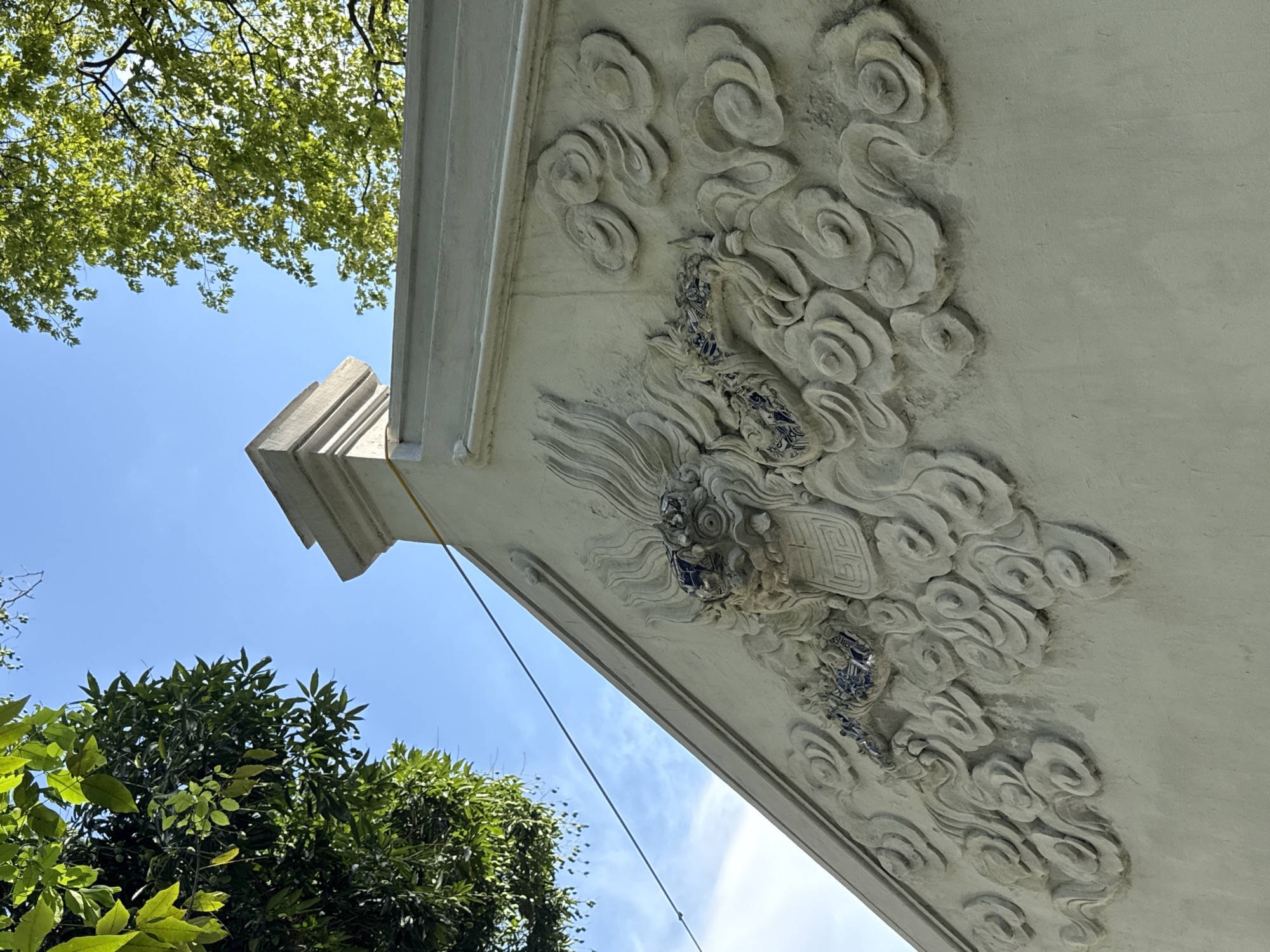The image depicts a low-angle view of a section of ornate stonework against a backdrop of trees and blue sky. The stonework is off-white and features raised relief carvings of swirling clouds and stylized dragon-like figures with blue tile accents. The stonework appears to be part of a building or architectural structure. The trees in the background have lush green foliage, and the sky is a clear blue with wisps of clouds.
Based on the architectural style and ornamentation, the scene is likely located within the city of Hà Nội, Vietnam.