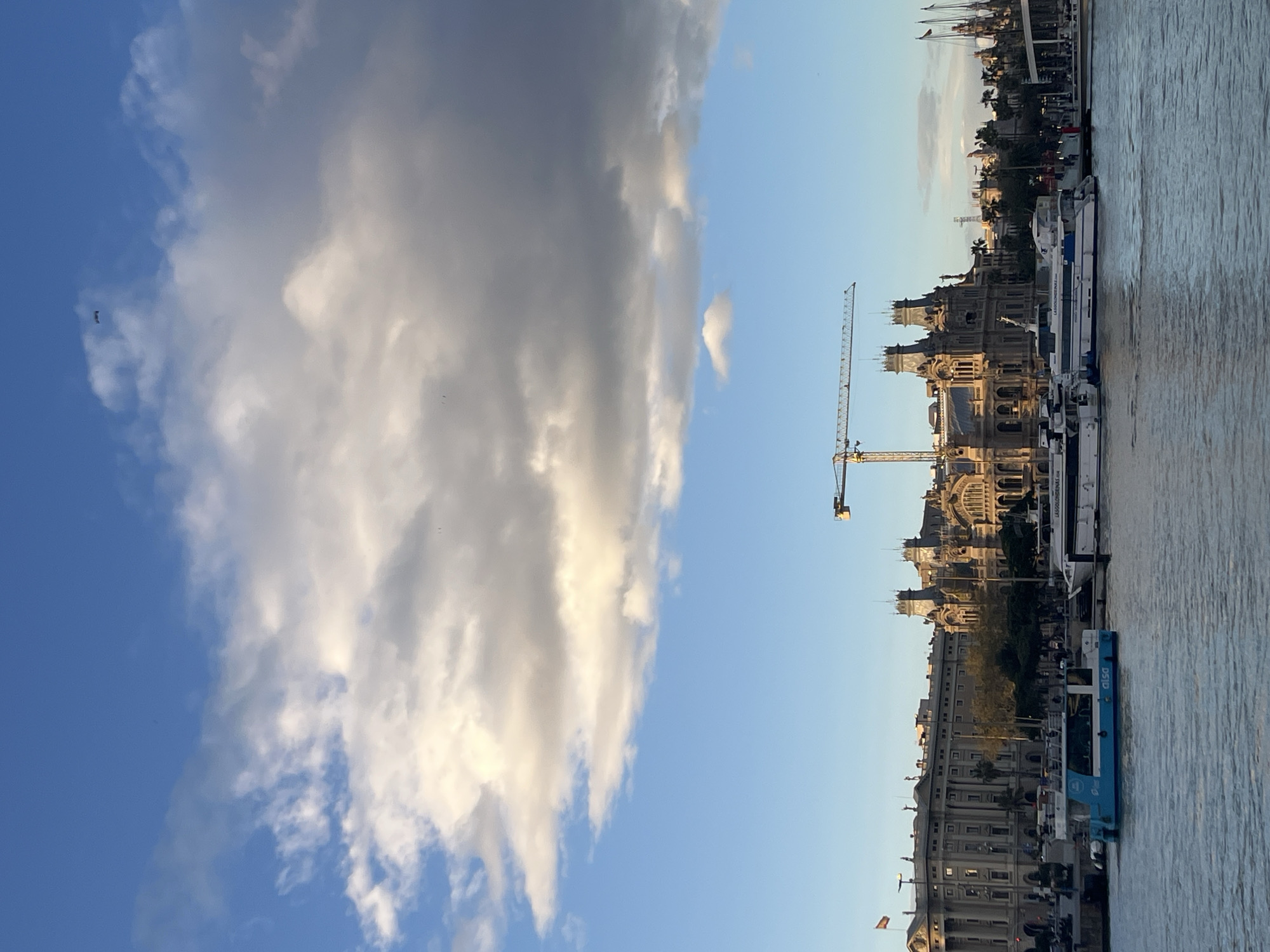 The image shows the city of Barcelona, Spain, on a bright day with blue skies and large white and gray clouds. The camera faces inland from the waterfront, capturing architectural details of buildings, including what appears to be the Port Authority building. A crane is visible near the buildings. Several boats are docked, including what appears to be the Las Golondrinas ferry. A blue and white bus is also docked. The photo is taken during the day with natural sunlight.