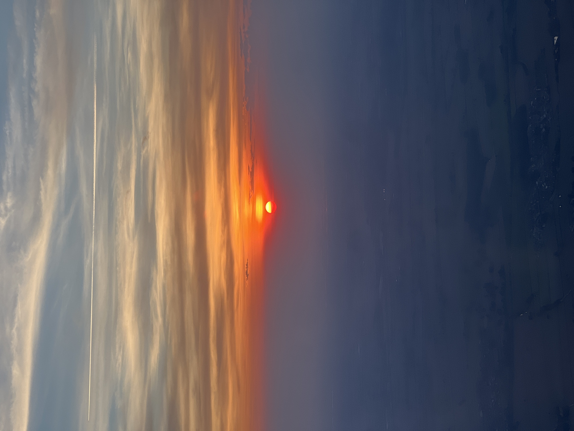 The image depicts a sunset in what appears to be a rural or semi-rural area. A bright, intensely red sun is partially obscured by a band of orange-tinted clouds. To the left of the sun, lighter-colored clouds stretch across a blue sky, marked by a single, straight contrail from an airplane. To the right of the sun and clouds, the sky transitions to a deep blue, suggesting either the onset of night or the effect of atmospheric perspective. There is no evidence of people, subjects, or objects, this image is the environment of the city of Spirago, Italy. The image is taken from a low angle, close to the ground.