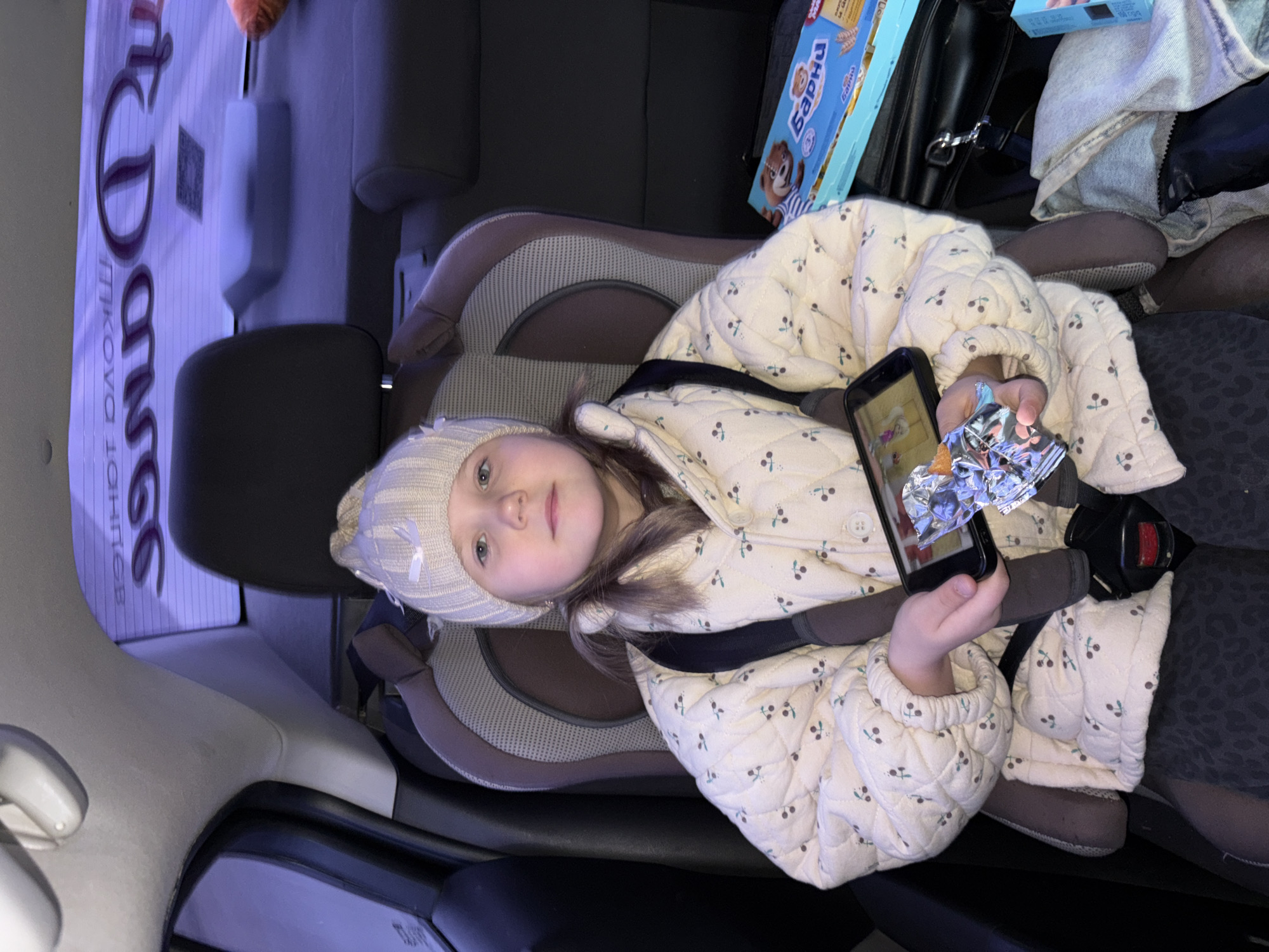 A young girl is sitting in a car seat in what appears to be the interior of a car. She is wearing a knitted hat and a patterned jacket. She is holding a smartphone in her hands, displaying an image on the screen, and in the other hand, she holds a foil package. The car interior includes seats, a box of "Barni" cakes, and possibly other belongings in the background. A sign for "My Donuts" is visible outside the car window, along with Cyrillic text that includes the location "Chaikovsky, Russia." She is secured with the car seat harness.