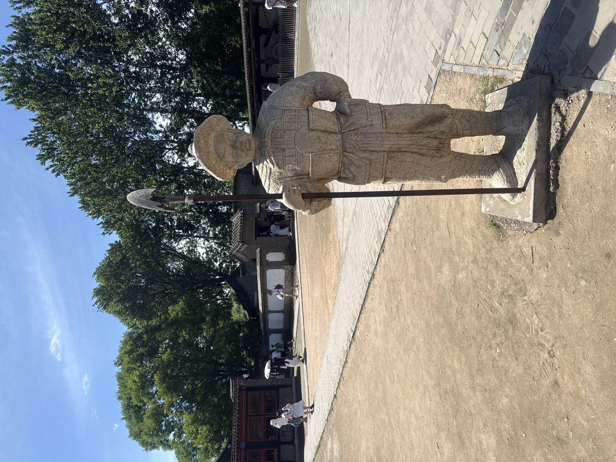 A stone statue of a warrior stands in the foreground, holding a spear. The statue is wearing a hat and armor, and its hands are on its hips. In the background, several people are walking around a courtyard with a paved walkway. There are buildings and trees in the distance. The scene is outdoors, under a clear blue sky with a few scattered clouds. This image was taken at the Shanhaiguan Great Wall Scenic Area in Qinhuangdao, China.