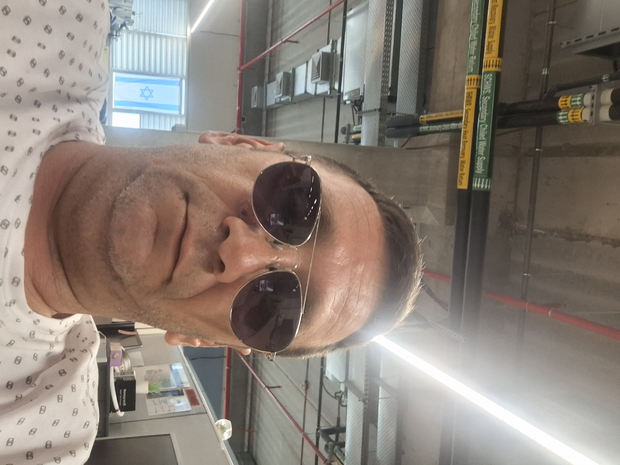 The image is a selfie taken indoors, likely in an office environment. The subject, a man wearing sunglasses and a patterned white shirt, is in the foreground. Behind him, various office elements are visible, including cubicle walls, a computer monitor, and what appears to be an Israeli flag hanging in a window. Overhead, industrial-style piping and lighting fixtures are present, consistent with a building's infrastructure. The scene appears to be in Kiryat Gat, Israel.