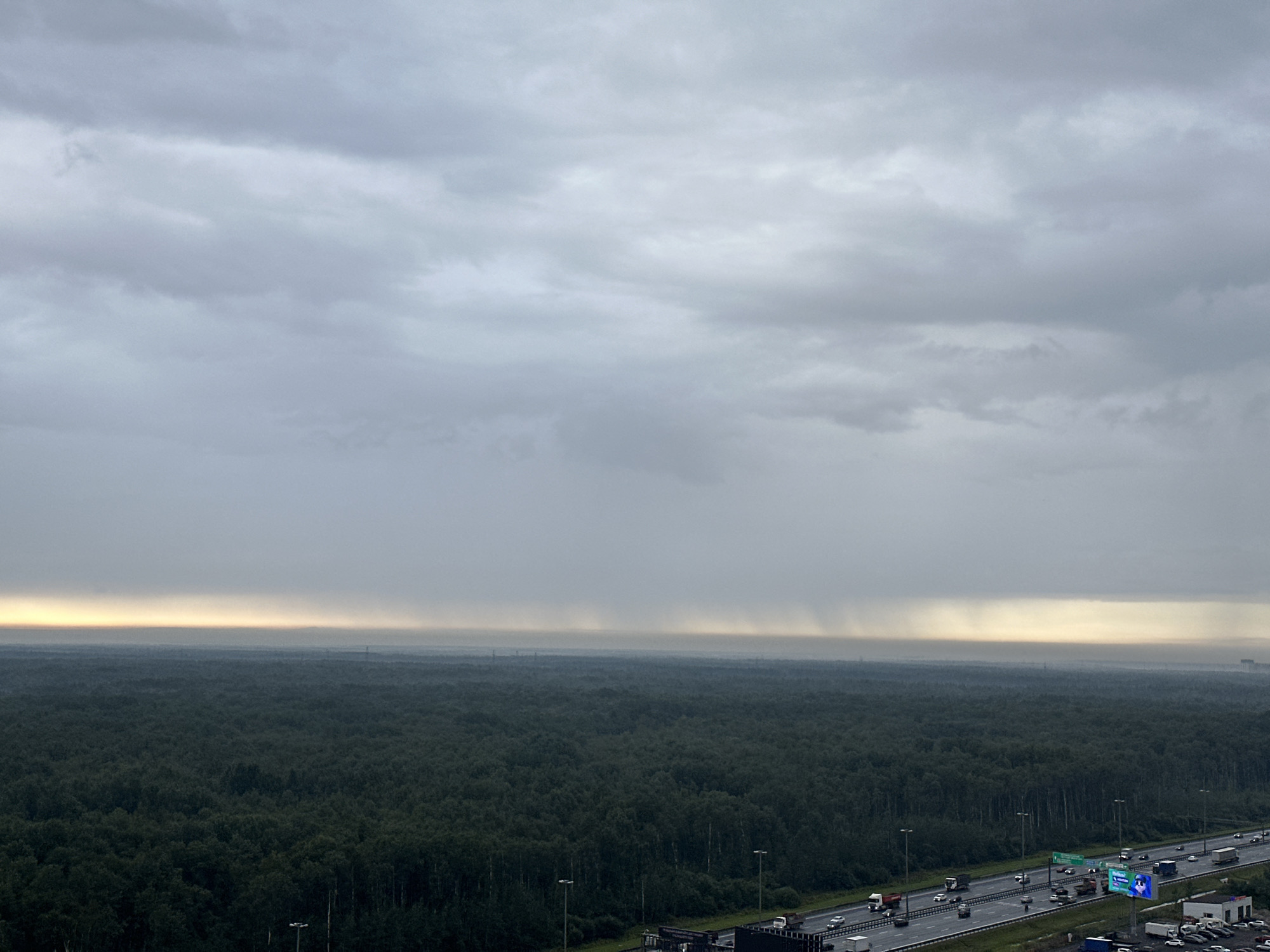 The image captures a wide-angle view of a dense, dark green forest stretching to the horizon under a heavily overcast, grey sky. A thin band of pale yellow light is visible along the horizon line, suggesting the presence of the sun behind the thick clouds or a very distant sunrise/sunset. Vertical streaks of what appears to be rain are visible in the distance against this bright horizon. In the foreground, a multi-lane highway with moderate traffic runs diagonally from the bottom right to the center. Vehicles, including cars and trucks, are moving in both directions on the highway. Streetlights are positioned along the side of the road. A large billboard with a blue and white advertisement is also visible. The overall atmosphere is somber and hints at impending or ongoing rain. This scene is located in the city of Kudrovo, Russia, as indicated by the presence of a highway and the landscape characteristic of the region.