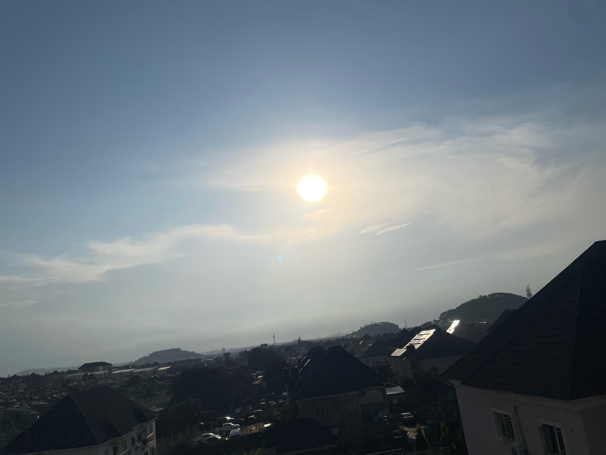 The image shows a panoramic view of a residential area in Abuja, Nigeria, under a bright, partially clouded sky. The sun, a prominent bright orb, is positioned in the upper center of the frame, casting a bright glare that washes out some of the details in the sky and distance. The foreground is dominated by the dark, angled roofs of several modern houses, suggesting a relatively affluent neighborhood. Beyond these houses, the cityscape extends into the middle ground, with more buildings, some appearing to be smaller or older structures, interspersed with trees. The background features rolling hills, characteristic of Abuja's topography. A few vehicles are visible on roads within the settlement. The overall impression is of a densely populated urban environment situated in a hilly landscape, bathed in the light of a sunny day.