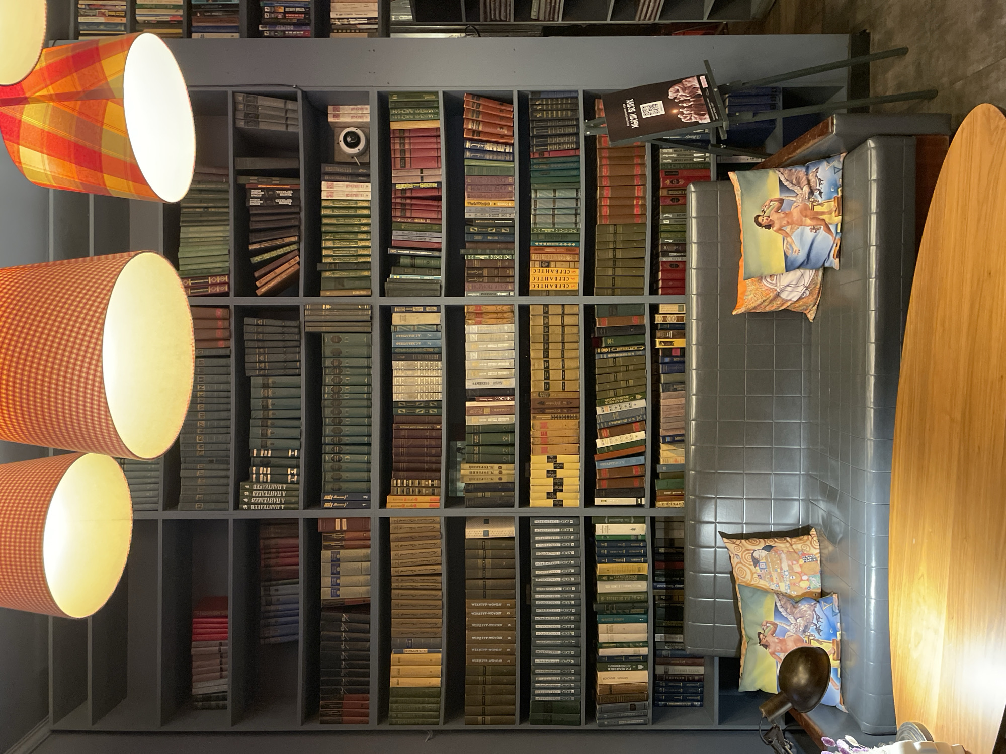 Here is a description of the image:
The image presents an indoor setting featuring a large, gray bookshelf filled with numerous books of varying sizes and colors. The books are arranged vertically in neat rows within the shelves. Adjacent to the bookshelf is a gray couch or seating area with two decorative pillows, one of which has a design resembling a Klimt painting, and the other depicts a historical scene with a person. A light wooden table is positioned to the right of the seating area. Above, three decorative lamps with colorful shades in orange and plaid patterns are suspended. The lighting illuminates the scene, enhancing the details of the books and furniture. The perspective appears to be from above, looking down onto the scene. Based on the presence of books written in Russian, and the sign "Мысли вслух" (Thoughts Aloud) near the couch, this image likely depicts a cozy corner in Saint Petersburg, Russia, specifically a library or reading room.