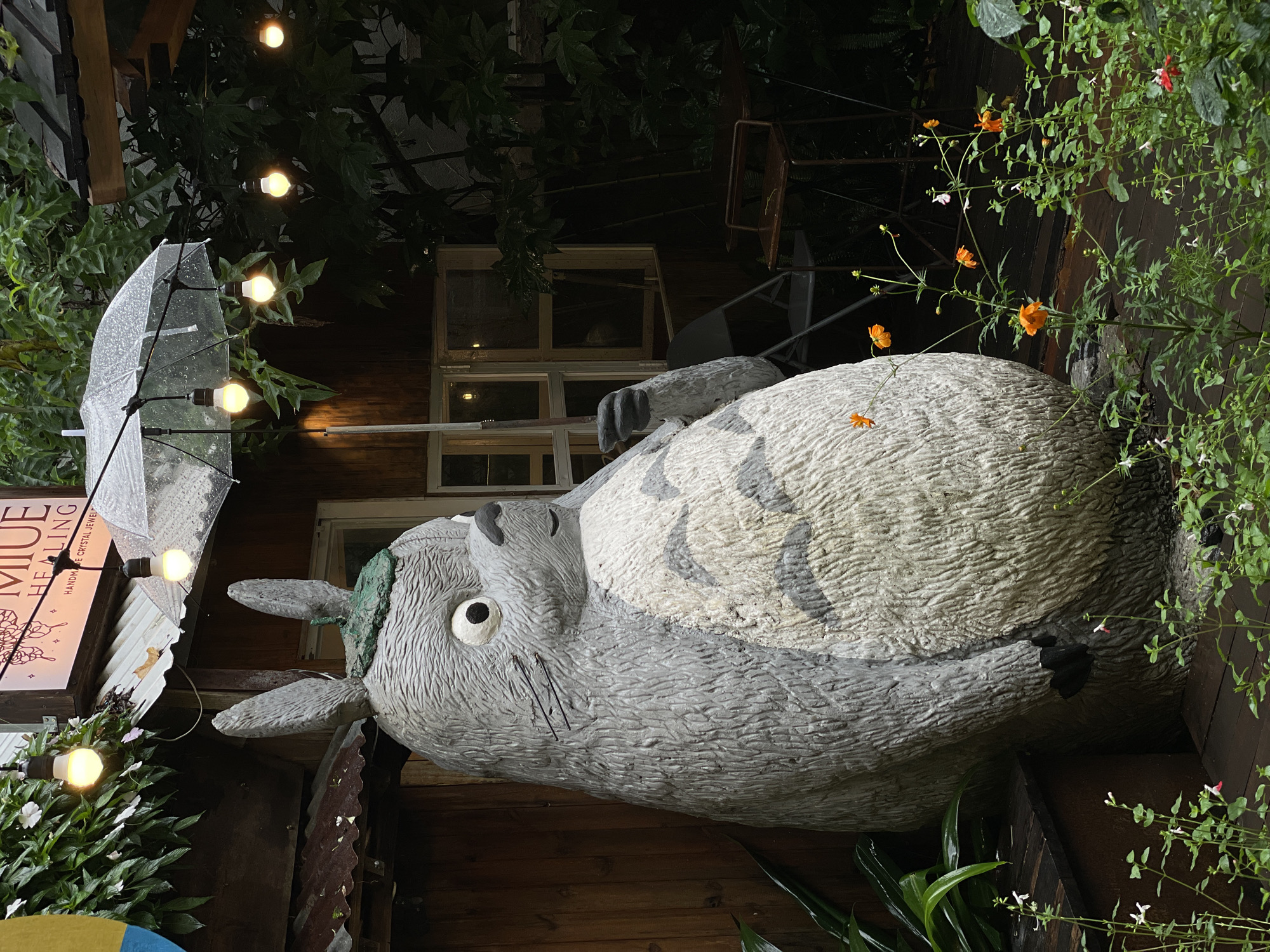 The image depicts a large, concrete Totoro statue holding a clear umbrella. Totoro's head is slightly turned towards the left. A sign for "Miue Healing" is visible to the left. The location is in Lâm Viên - Đà Lạt, Vietnam. The setting appears to be a garden or outdoor area adjacent to a building with wooden siding and windows. Strings of lights are strung around the area, and various plants and flowers surround the statue.