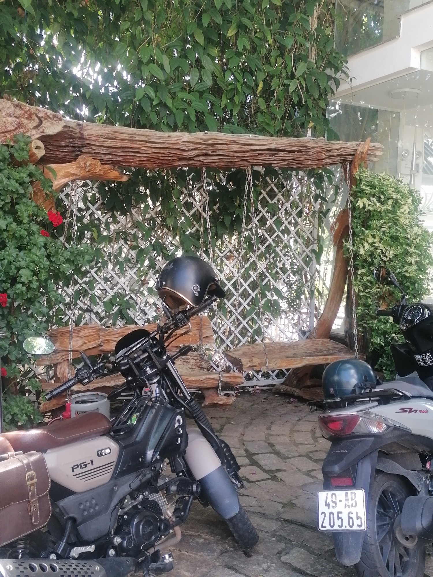 The image depicts a scene with two motorcycles parked outdoors. The foreground features a Yamaha PG-1 motorcycle, distinguished by its brown leather saddlebag and a black helmet resting on its handlebars. To the right, a white Honda scooter is visible, with another helmet, this one blue, placed on its rear rack. Both vehicles are parked on a cobbled surface, suggesting an outdoor setting.
Behind the motorcycles, a rustic wooden swing hangs from chains, suspended from a rough-hewn wooden beam. The swing is set against a backdrop of lush green foliage and a white lattice fence. Red flowers are also present among the greenery, adding a touch of color to the scene. The presence of a modern building with glass elements is partially visible in the background, hinting at a location that combines natural elements with contemporary architecture.
While the exact location within Da Lat is not explicitly stated in the image, the overall aesthetic—the blend of lush greenery, rustic wood, and the presence of motorcycles—is characteristic of many cafes, guesthouses, or residential areas in the city, known for its charming, often nature-integrated, outdoor spaces. The license plate on the Honda scooter, "49-AB 205.65," indicates its registration in Lam Dong province, which includes Da Lat, further solidifying the geographical context.