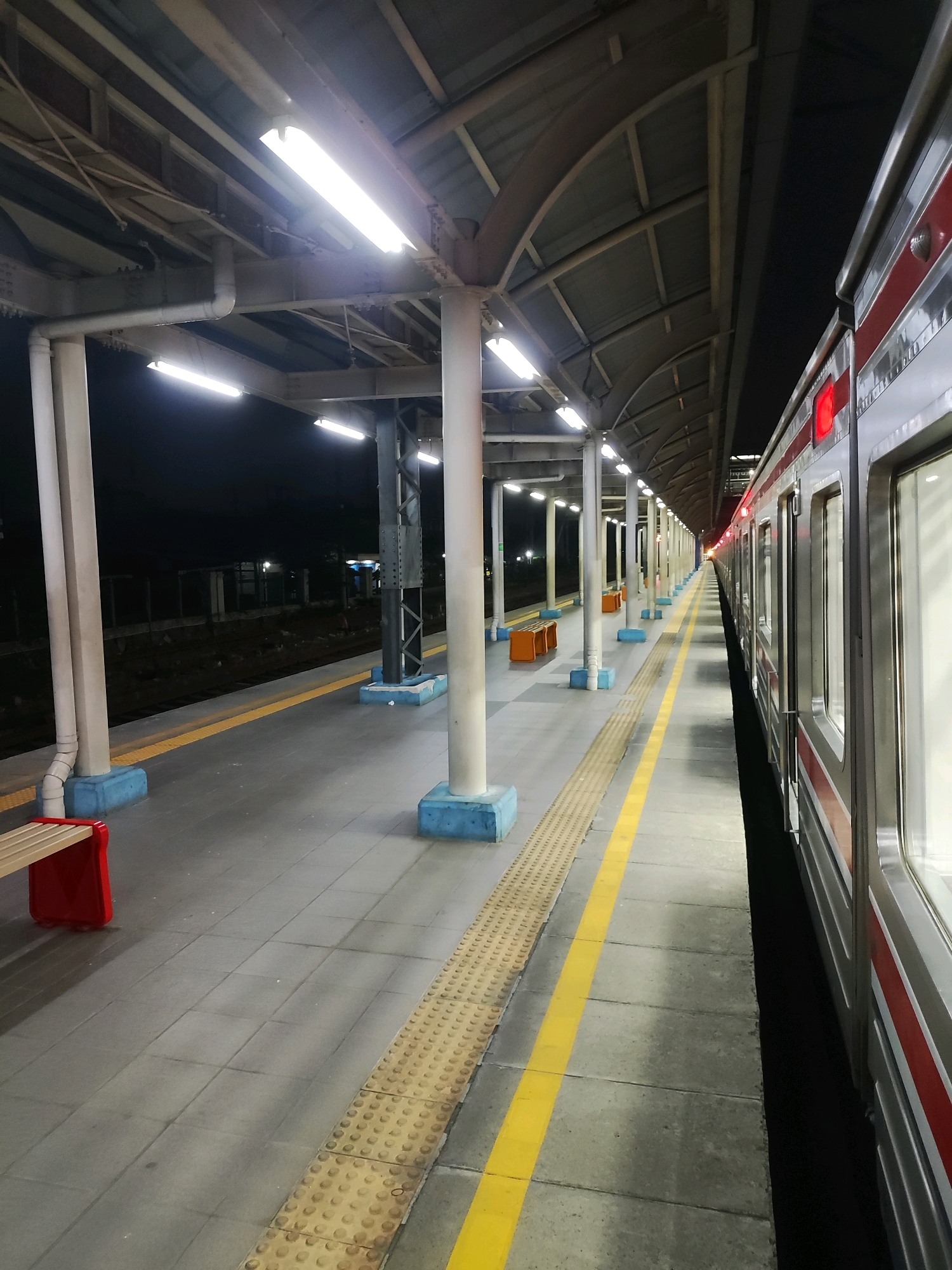The image depicts a train station platform at night in Tambun, Indonesia. A train with silver and red livery is stopped alongside the platform. The platform is paved with grey tiles and features a tactile paving strip for visually impaired passengers, along with a bright yellow line demarcating the edge. Several white pillars with blue bases support a corrugated metal roof that extends over the platform, illuminated by fluorescent lights. Two orange benches are visible further down the platform, and a red-seated bench is in the foreground. The background shows dark, unlit tracks and the silhouette of distant structures. The scene is empty of people.