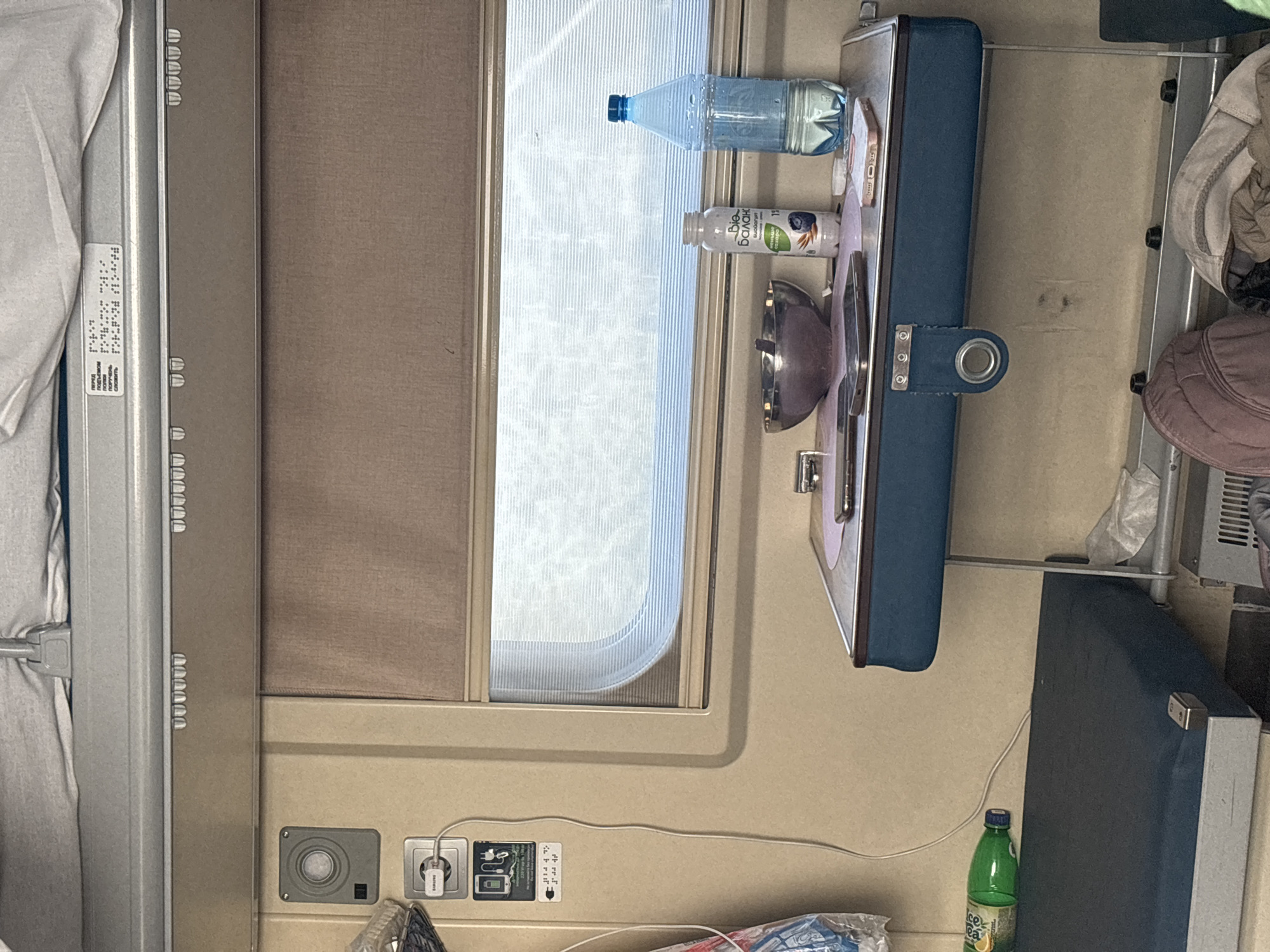 The image displays the interior of a train cabin, specifically a berth or compartment, situated in or en route to/from Samarka (Samara), Russia, as indicated by the prompt and the presence of Russian-language labels within the cabin.
The cabin walls are uniformly light beige. An upper bunk or luggage rack, covered with light grey fabric, is visible at the top of the frame. Below this, a grey metallic strip runs horizontally, featuring small, evenly spaced protrusions along its length. A label affixed to the overhead structure contains text in Russian and Braille, with visible phrases including "ПЕРЕД ОТПРАВЛЕНИЕМ ПОЕЗДА" (BEFORE TRAIN DEPARTURE).
On the left wall, a grey circular fixture, possibly a light or sensor, is mounted above a standard power outlet. A Samsung brand wall charger is plugged into this outlet, with a white charging cable extending downwards. Below the outlet, a sticker illustrates mobile device charging icons and includes Braille text.
A window with a drawn brown horizontal blind provides a diffused view of a bright exterior. Below the window, a folding table, upholstered in dark blue with light brown accents, is extended horizontally. The table surface is partially covered by a light pink mat. Items arranged on the table include: a metallic bowl containing dark, possibly food-related, contents and a spoon; a clear plastic bottle of "Bio-Баланс" (Bio-Balance) yogurt with a blueberry graphic; a clear plastic water bottle with a blue cap, partially filled; a dark-colored smartphone resting face down; and a lighter-colored smartphone with a patterned case, resting face up. A small piece of crumpled white tissue is also present near the water bottle.
Below the table, a lower bunk or seat with dark blue upholstery is visible. A green plastic bottle, bearing a visible "Sprite" logo, lies horizontally on this bunk. Beneath the table, on the floor, a light brown backpack and a crumpled light brown garment or blanket are positioned, along with a partially visible white plastic bag. No individuals are present in the scene.
