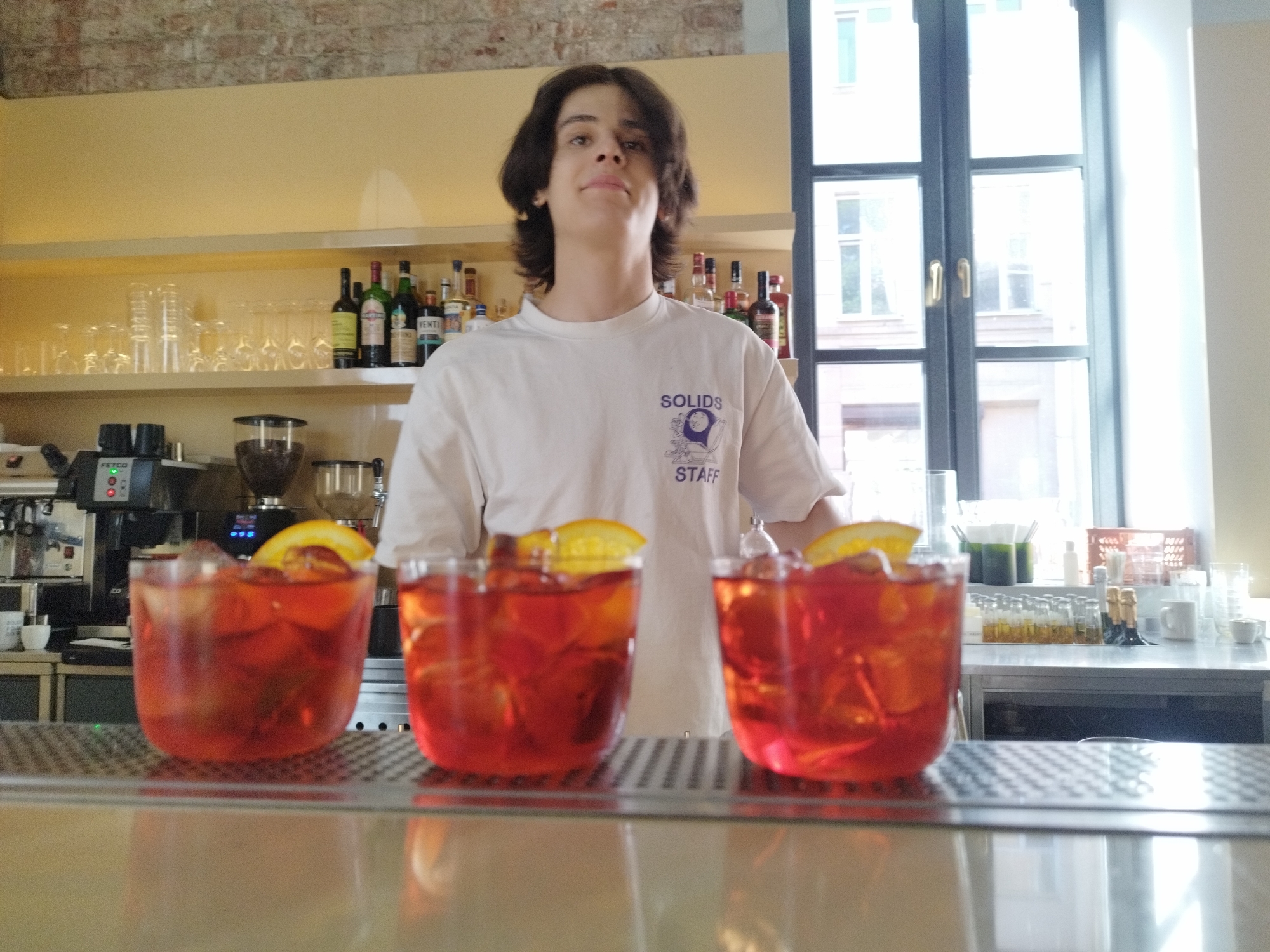 A young person stands behind a bar, wearing a white t-shirt with the text "SOLIDS STAFF" and a graphic. In front of them, three glasses of red cocktails with orange slices are prominently displayed on the bar top. Behind the cocktails, a variety of bottles, glasses, and bar equipment are visible on shelves and counters. The setting appears to be a bar or restaurant, with a brick wall in the background and large windows to the right, suggesting daylight. The person has a neutral expression. The scene is well-lit, with light reflecting off the bar surface.