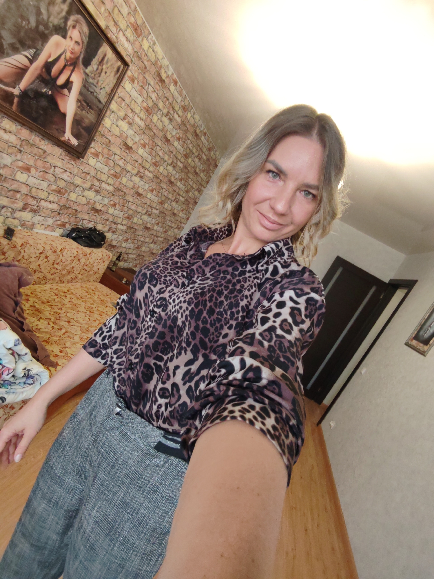 A woman is captured in a selfie indoors, likely within a bedroom or living area in Petropavlovsk-Kamchatsky, Russia. She is smiling faintly at the camera, holding it herself with an extended arm. She has light, wavy hair and is wearing a dark brown and black leopard print long-sleeved shirt tucked into gray plaid-patterned trousers.
The background reveals a room with distinctive features. A prominent wall behind her has a textured brick-patterned wallpaper. On this wall, a large, framed picture hangs, depicting a woman in a black bikini posing on a rocky or wet surface. To the left, part of a bed or sofa with a light brown, floral-patterned cover is visible, with a dark bag or piece of clothing resting on it. The floor is covered with light wooden planks.
In the right background, a dark brown door with vertical panels is partially visible against a plain, light-colored wall. A strong light source, possibly an overhead lamp or window, is positioned high above the woman's head, casting a bright glow across the upper part of the image. The time of day is not discernible, but the room is well-lit. There is no visible text in the image. The overall impression is a casual, personal moment captured in a homely setting.