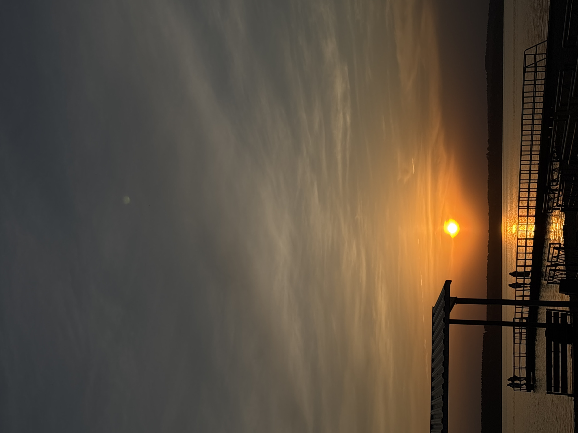 The image depicts a sunset at the waterfront. The sun, appearing as a bright orange orb, is positioned near the horizon, reflecting on the water's surface. The sky exhibits a gradient of colors, transitioning from a darker grey on the left to lighter orange hues towards the sun. A structure or pier with a grid-like pattern is visible on the right side of the image, partially silhouetted against the bright sky. The location is in the city of Perm, Russia. The image is oriented vertically.