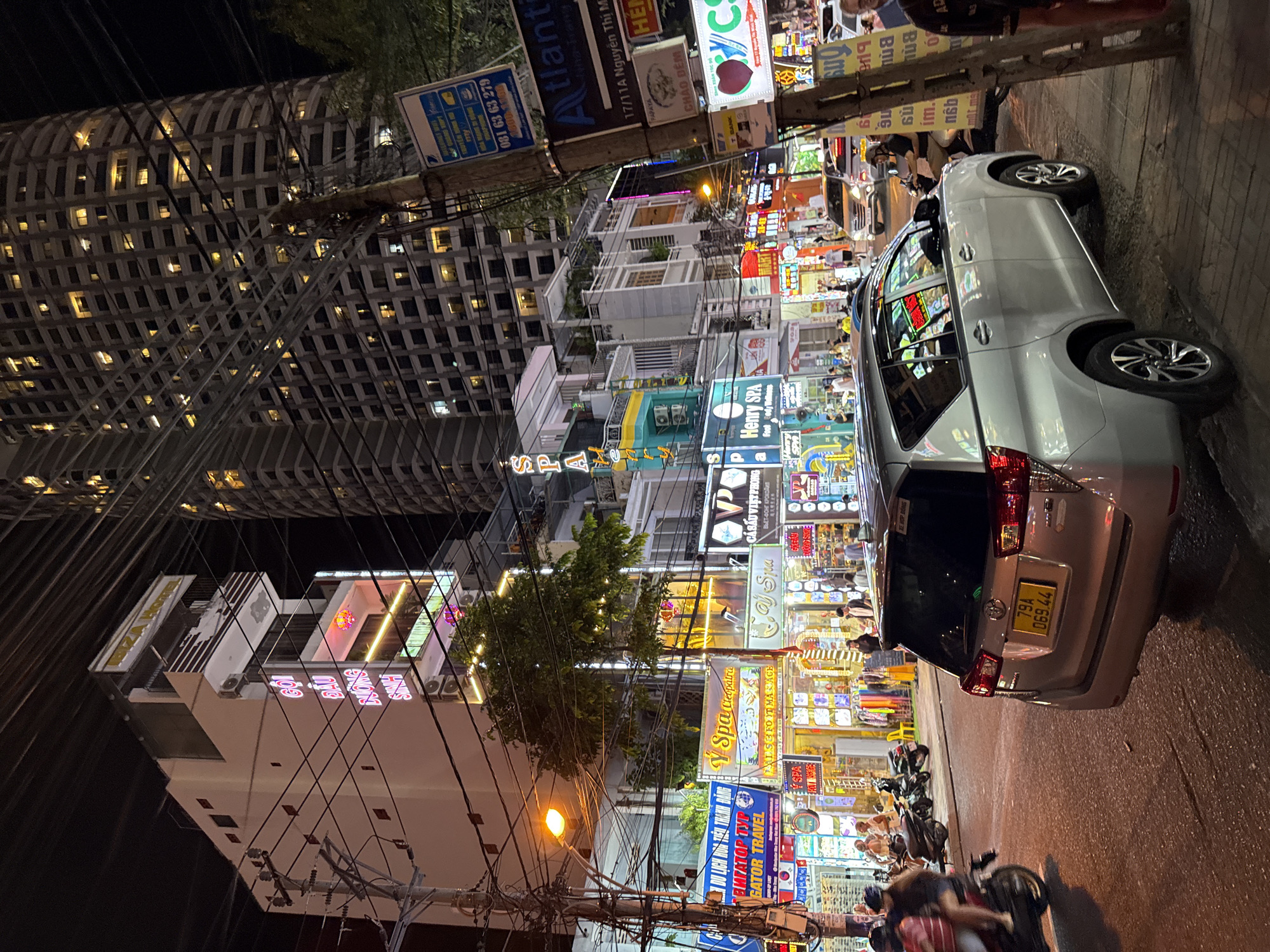 The image depicts a street scene at night in Nha Trang, Vietnam, specifically on a street likely to be tourist-oriented given the numerous illuminated signs for hotels, spas, and businesses. In the foreground, a silver Toyota Innova SUV is parked on the right side of the road. The vehicle's license plate is clearly visible, reading "79A 069.44". To the left of the SUV, a motorcycle carrying two people, a driver and a passenger, is in motion, heading away from the camera.
The street is lined with buildings of varying heights. On the left, a multi-story building displays prominent neon signs, including "IBIZA HOTEL" at the top and other signs with Vietnamese text, some illuminated in pink and green. Further down the street, a row of shops and businesses are visible, identified by illuminated signs for "Y Spa Cleopatra," "Henry SPA," and various other services. Some of these establishments have large windows displaying goods.
In the background, a very tall, modern high-rise building with many illuminated windows dominates the skyline, suggesting a significant urban development. The street is cluttered with a dense network of electrical wires suspended from utility poles, a common sight in many Asian cities. Several other vehicles and pedestrians are present on the street, with people walking along the sidewalks and some standing in front of the businesses. The overall atmosphere is one of a busy, commercial street at night, illuminated by a mix of streetlights and neon signage.