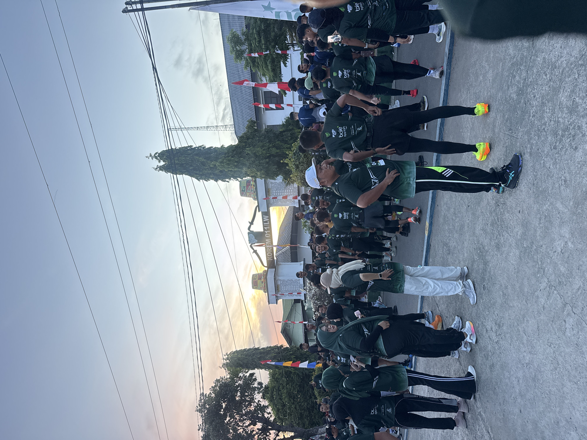In the city of Lhokseumawe, Indonesia, a group of people are gathered outdoors in front of a building with the text "KOREM 011/LW" above its entrance. The sky is a gradient of pale blue to pink, suggesting it is either early morning or late evening. The individuals, many wearing matching dark green t-shirts and black pants, appear to be participating in or preparing for an event, possibly a run, given the athletic attire and the presence of a few flags. Some people are wearing headscarves, indicating a mix of genders and possibly cultural attire. The ground is paved with concrete. The overall scene depicts a community gathering or event taking place in a public space.
