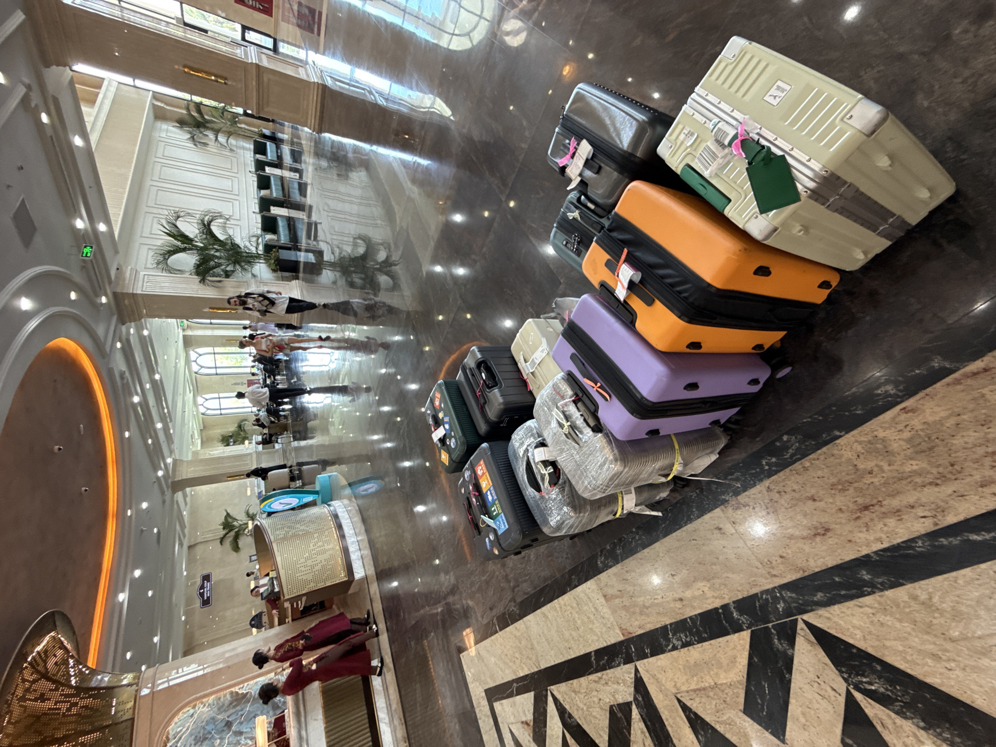 The image depicts a luggage pile in a hotel lobby. There are various colored suitcases and wrapped bags stacked together. The lobby has reflective, dark-colored marble floors with patterned inlays near the luggage. In the background, there is a reception desk with two staff members, and the overall architecture suggests a luxurious hotel. The location is Nha Trang Ward, Vietnam. People can be seen walking towards the reception desk.