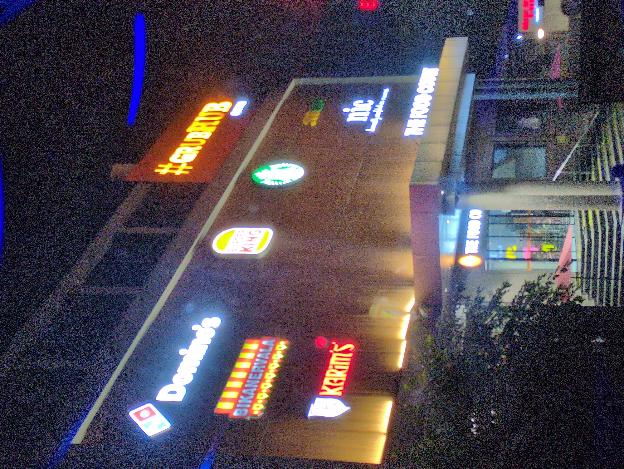 The image shows the exterior of a building at night, likely a commercial complex or mall, located in Una, India. The building is illuminated by numerous neon signs advertising various food establishments. Prominently visible are signs for Domino's, Bikanervala, Burger King, Starbucks, and Karim's. A sign with "#GRUBHUB" is also visible. Towards the bottom right, a sign reads "THE FOOD COURT". The architecture appears modern, with the signs mounted on the facade of the building. The overall atmosphere is vibrant due to the bright neon lights against the dark night sky. There are no people clearly visible in the frame. The ground level shows stairs leading up to the building entrance, and some greenery is present on the left side.