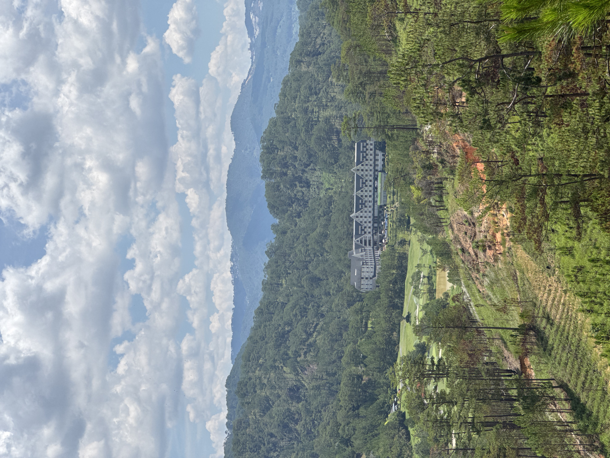 A picturesque landscape showcases the serene beauty of Hiep Thanh, Vietnam. In the foreground, a verdant hillside is adorned with tall, slender pine trees, their needles a vibrant shade of green. The landscape descends into a manicured golf course, its fairways and greens sharply contrasting with the rugged natural environment.
Nestled amidst the lush greenery is a contemporary hotel building. Its architecture is characterized by a series of gabled roofs and a neutral color palette. A row of parked vehicles hints at activity.
In the distance, rolling hills and mountains fade into a hazy blue, adding depth to the composition. The sky is filled with fluffy white clouds. The overall impression is one of tranquility and natural splendor.
