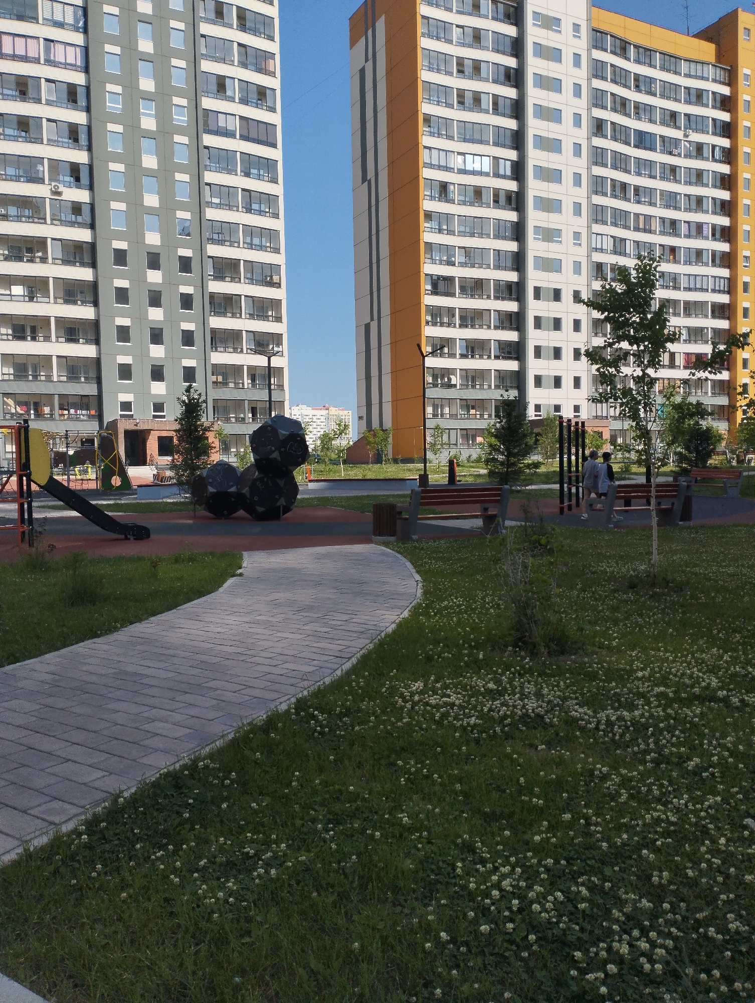 This image depicts a modern residential courtyard in Sadovyy, Russia. Two tall, multi-story apartment buildings dominate the background. The building on the left has a gray facade with numerous white-framed windows and balconies, while the building on the right features a striking orange and white exterior. In the foreground, a paved walkway curves through a grassy area dotted with small white flowers. Adjacent to the walkway is a children's playground with colorful equipment, including a slide and climbing structures. Several dark, sculptural elements resembling rocks are also present in the play area. Further back, benches and outdoor fitness equipment are visible. Two individuals are walking in the distance, towards the right side of the frame, near the benches. The scene is bathed in natural daylight under a clear blue sky.