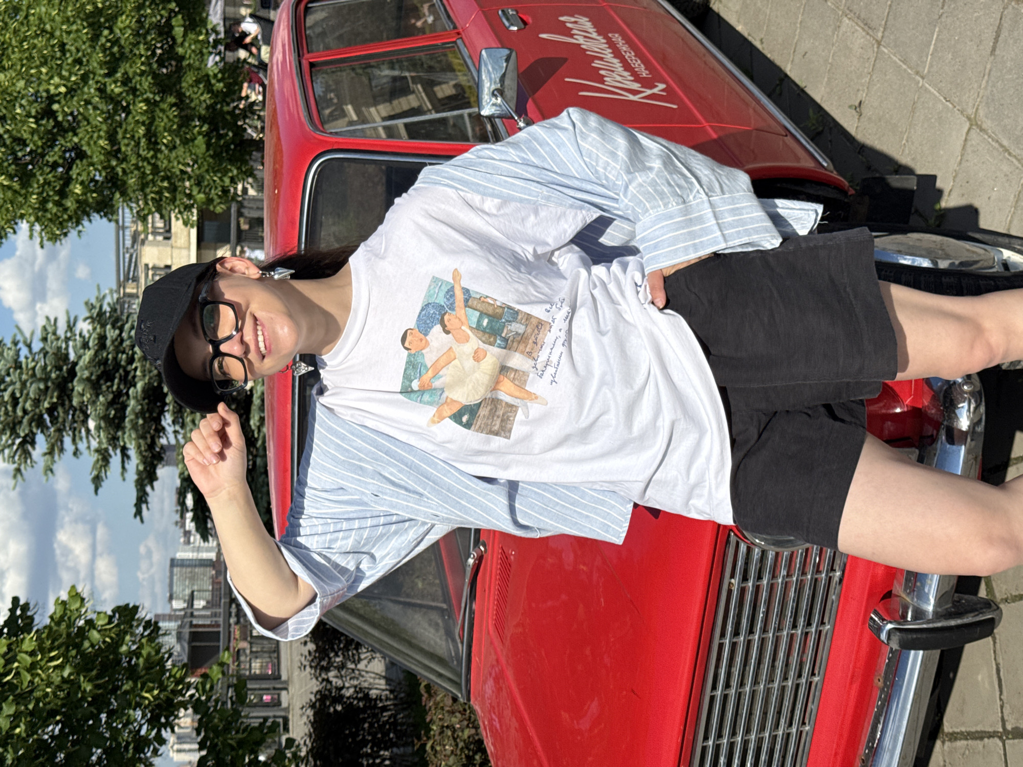 A person wearing a black baseball cap and glasses is standing in front of a red car, with greenery and buildings visible in the background, in what appears to be the city of Kazan, Russia. The person is wearing a white t-shirt with a graphic and a blue and white striped button-down shirt, along with black shorts. The red car has the word "KAZANSKAYA" written on it in white lettering. The image appears to be taken outdoors during the daytime, with a bright sky and sunlight casting shadows.