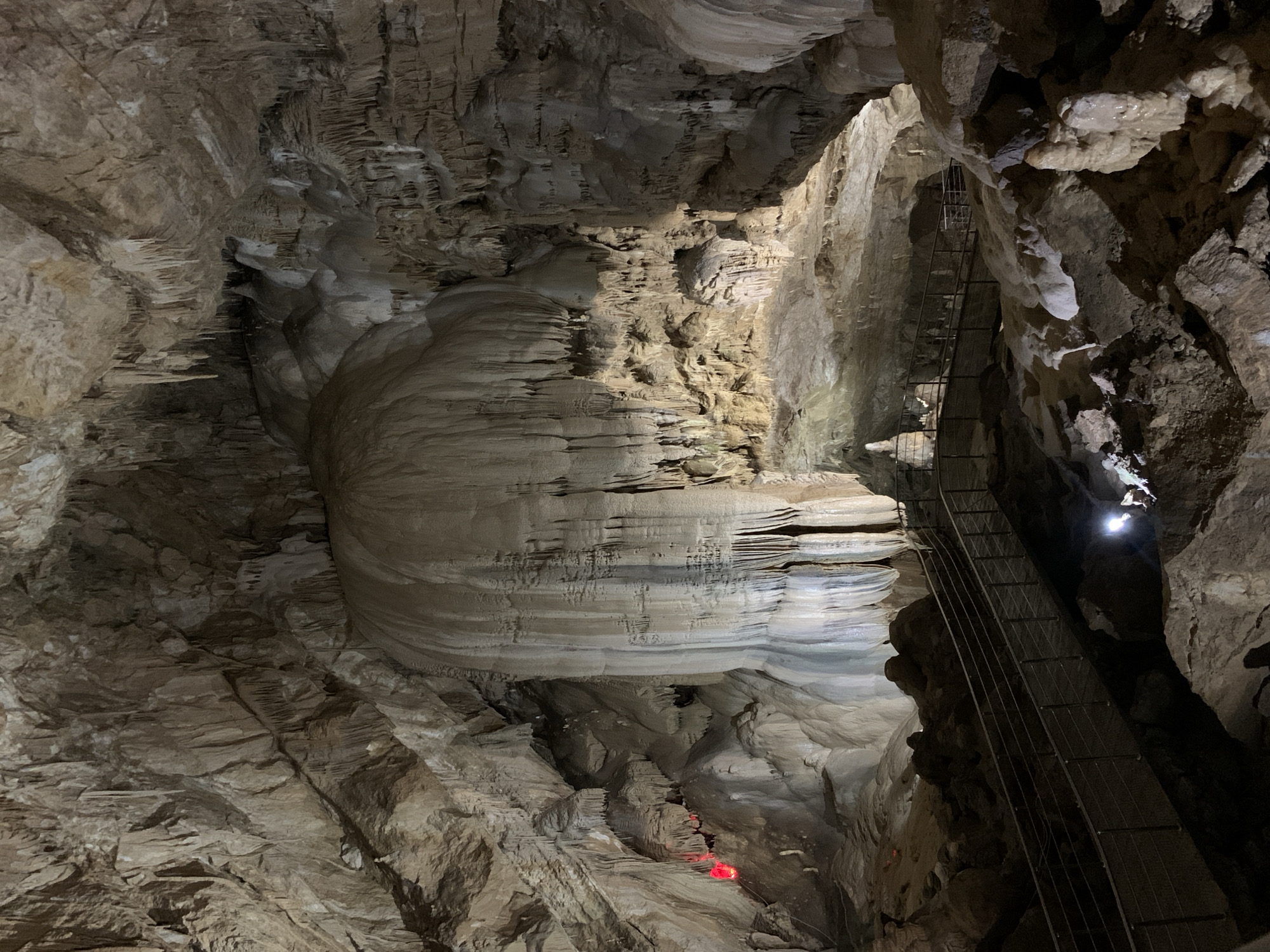The image depicts a cave interior, likely a tourist attraction, with prominent rock formations, stalactites, and stalagmites. A metal staircase ascends along one of the cave walls, possibly providing access for visitors. A small red light is visible at the bottom of the frame, potentially marking a path or hazard. A brighter white light illuminates an area higher up, possibly a natural opening or artificial lighting fixture. The environment is characterized by the textures and colors of natural stone, with varying shades of gray and brown. The location is purportedly in Inkiti, Abkhazia.