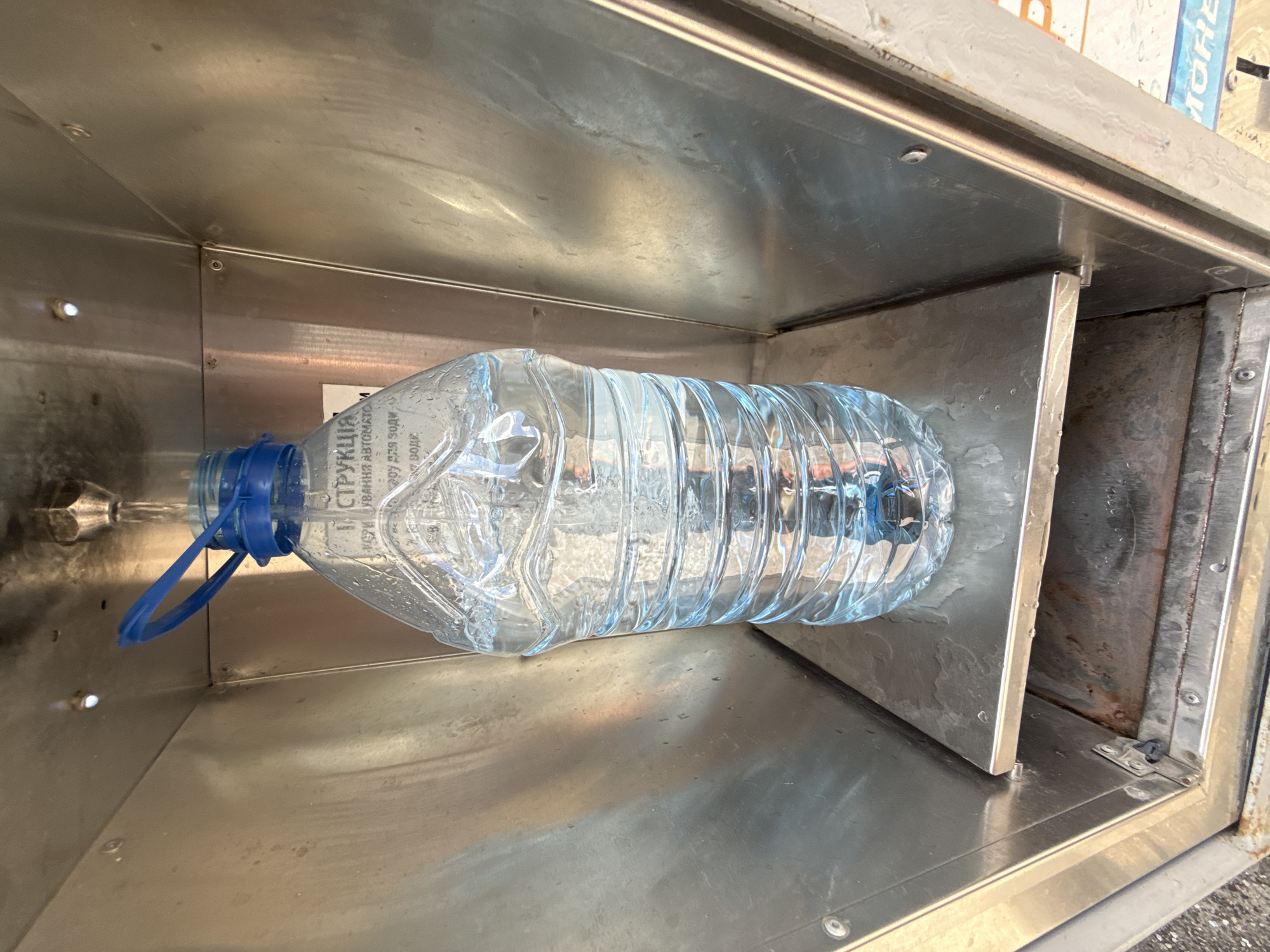 A plastic bottle of water is being filled from an overhead dispenser within a stainless steel kiosk. The bottle is partially full, with water visibly flowing into it. The kiosk appears to be outdoors, with a hint of pavement visible in the lower portion of the image. A label with Cyrillic text is attached to the bottle. The overall scene suggests a public water dispensing station, possibly in the city of Zaporizhzhia, Ukraine, given the context.