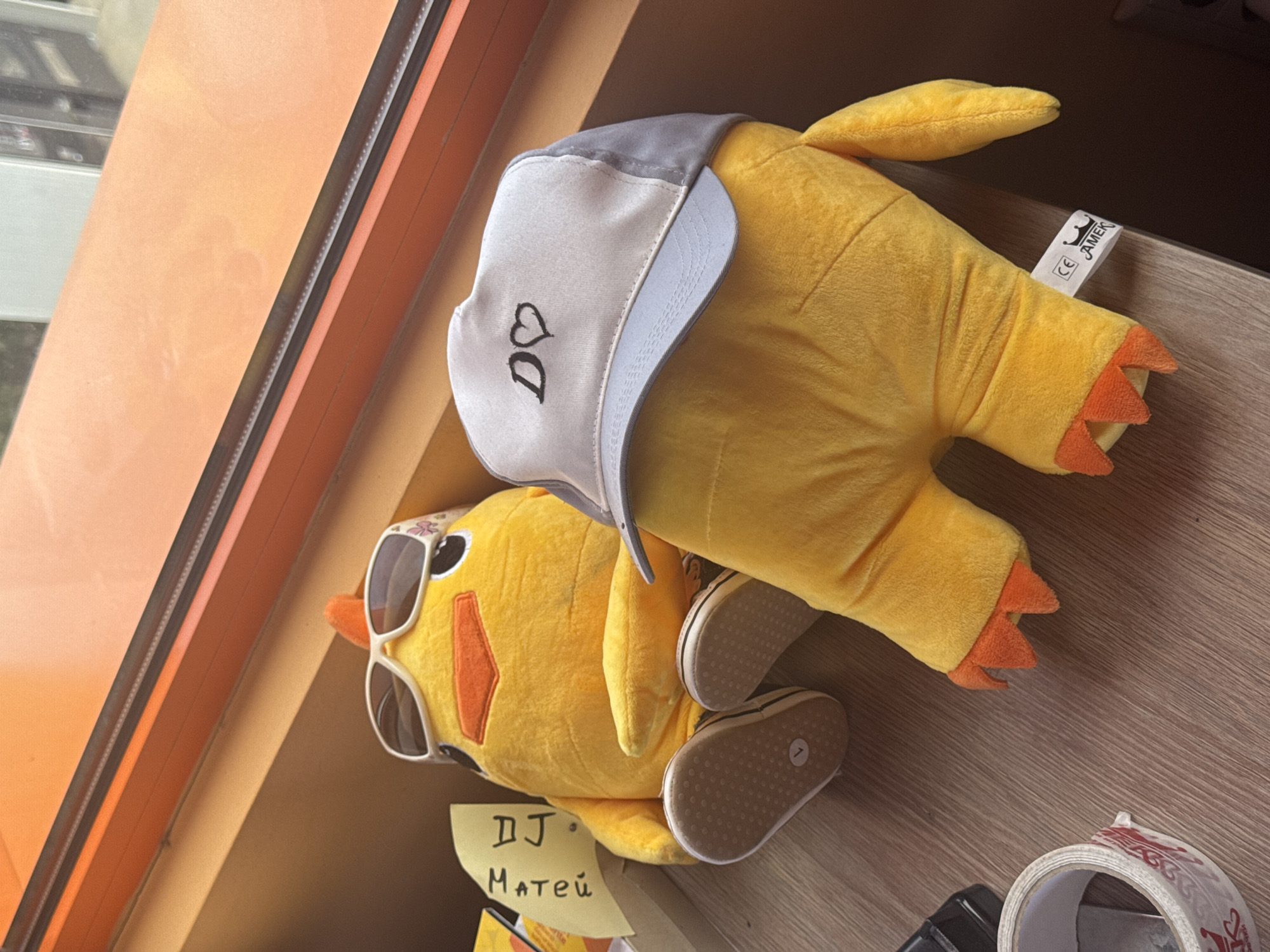 The image shows two stuffed yellow toys on a wooden surface, positioned near a window. One toy wears sunglasses and the other a grey and white cap. A yellow sticky note with the handwritten words "DJ Matei" is partially visible beneath the toys. A roll of clear tape, and a black object are present near the bottom edge of the frame. The location is the city of Unknown, Bulgaria.