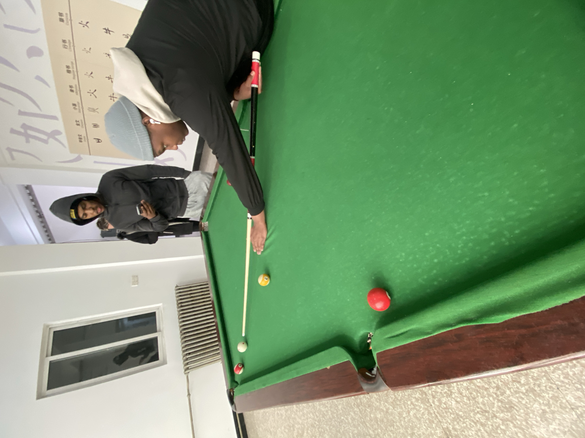 The image shows two individuals in an indoor setting, engaged in a game of pool on a green felt pool table. One person, wearing a grey beanie and a black and white hooded jacket, is leaning over the table, cue in hand, preparing to strike the cue ball. A second individual, standing behind, is wearing a black hooded jacket and holding a mobile phone in their hand.
The pool table has several balls on it: a white cue ball, a yellow ball, and two red balls. The table's wooden frame is visible, as is the green felt surface.
The setting appears to be a room with white walls. A radiator is visible on the wall, along with a window allowing a glimpse of the outside. The location is Zhongtun, China. Behind the two individuals, there's a framed piece of art or calligraphy with Chinese characters hanging on the wall.